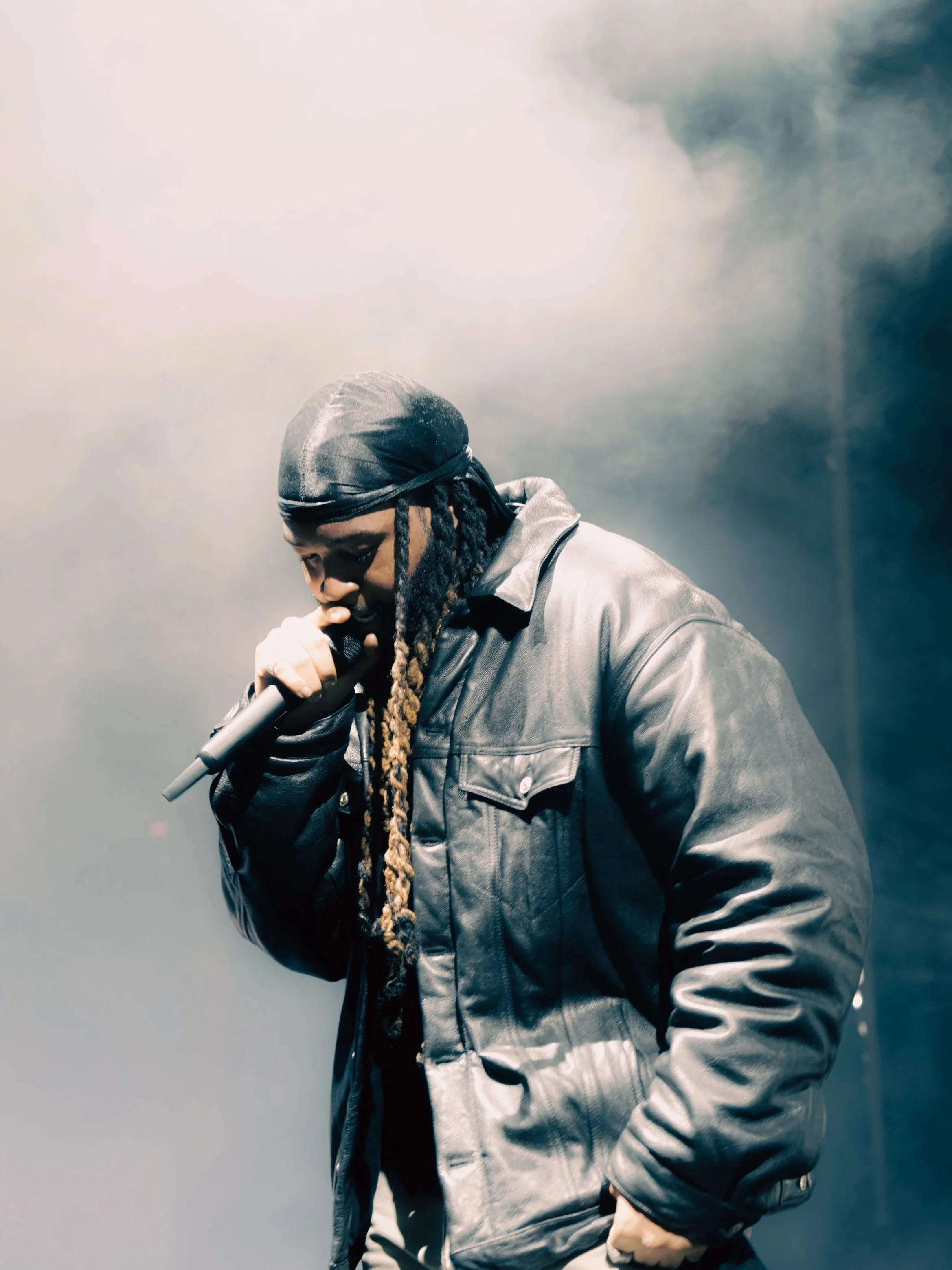 A man with long dreadlocks wearing a black leather jacket and a durag, holding a microphone and singing or rapping on stage.