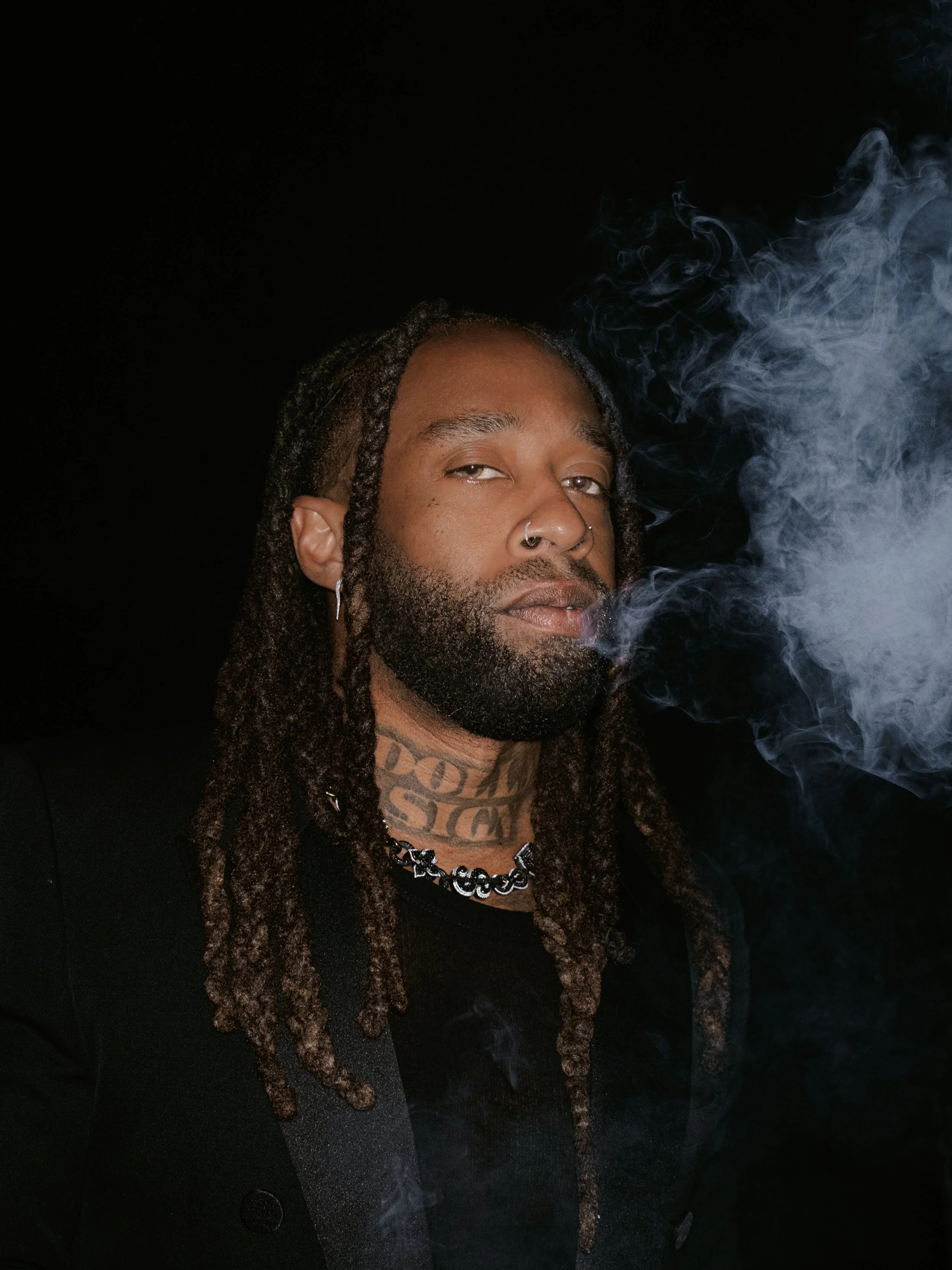 Photo of Ty Dolla $ign with dreadlocks, tattoos, and a nose ring smokes and exhales smoke against a dark background. By celebrity photographer.  Los Angeles