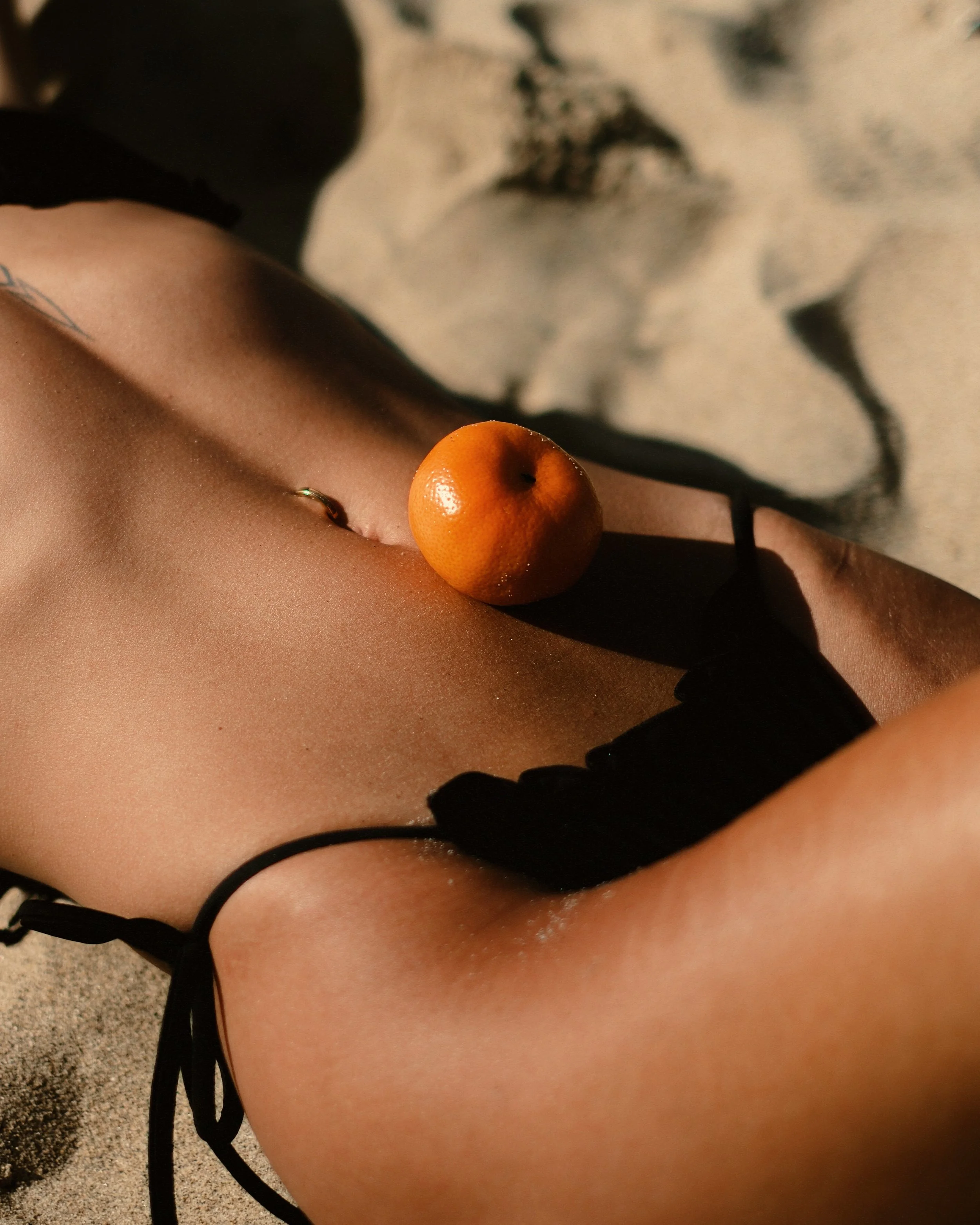 A close-up of a person's midsection on a sandy beach, with a small orange placed on their stomach and wearing a black bikini.