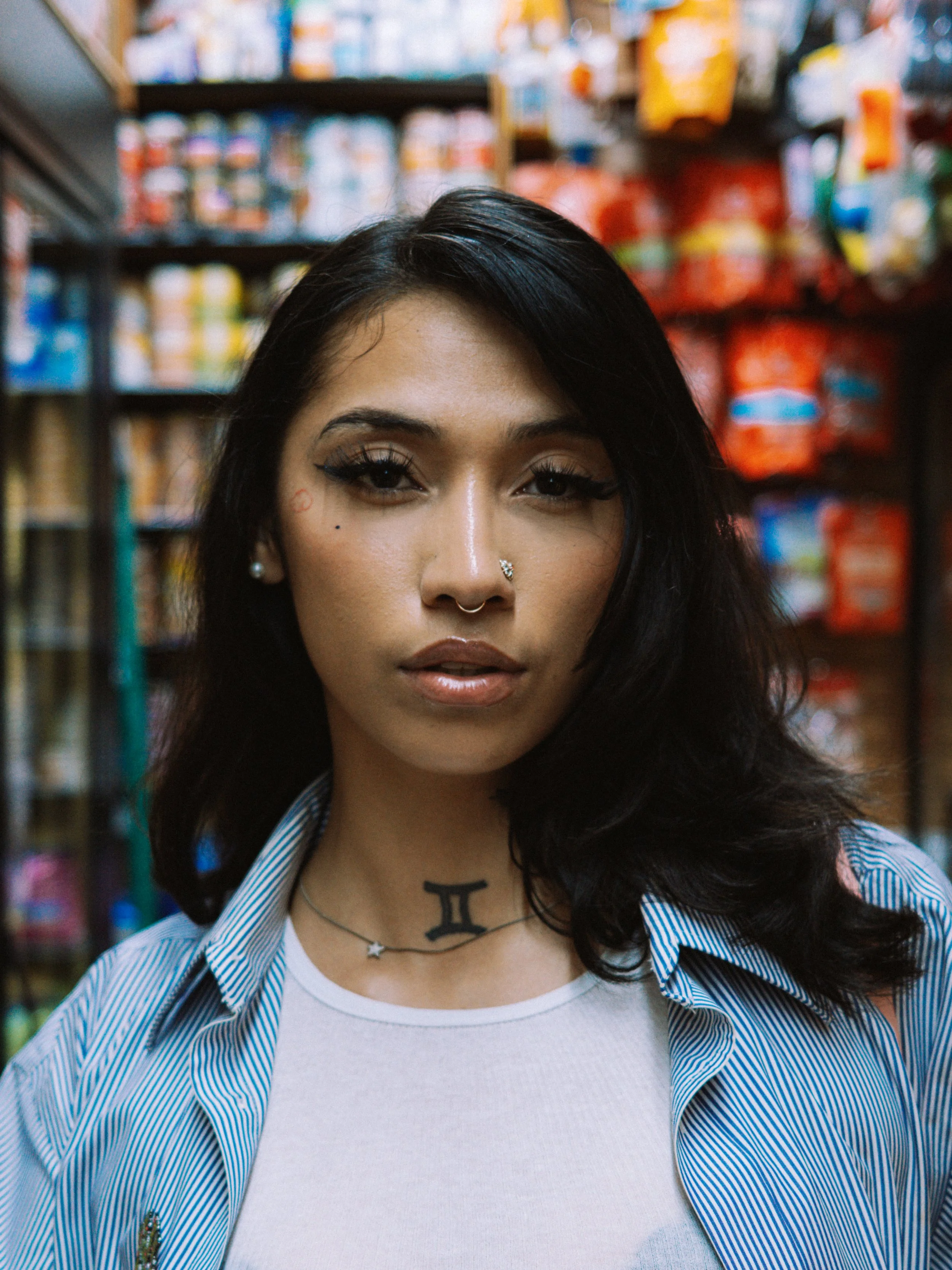 New York Rapper.  A young woman with dark hair and tattoos, standing in a grocery store aisle with snacks on shelves in the background.
