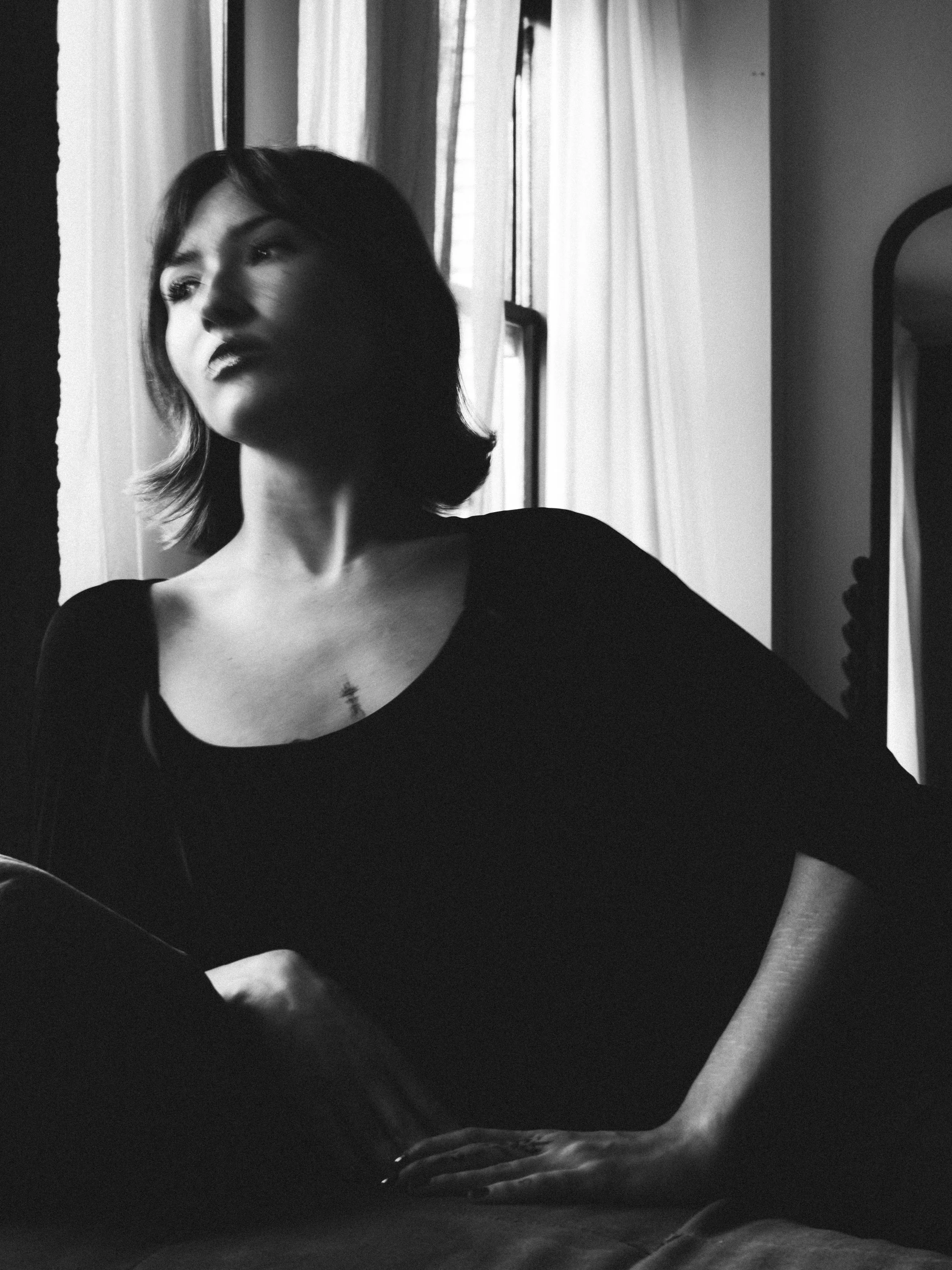 Los angeles model.  Black and white photo of a woman sitting near a window with curtains, looking thoughtfully into the distance.
