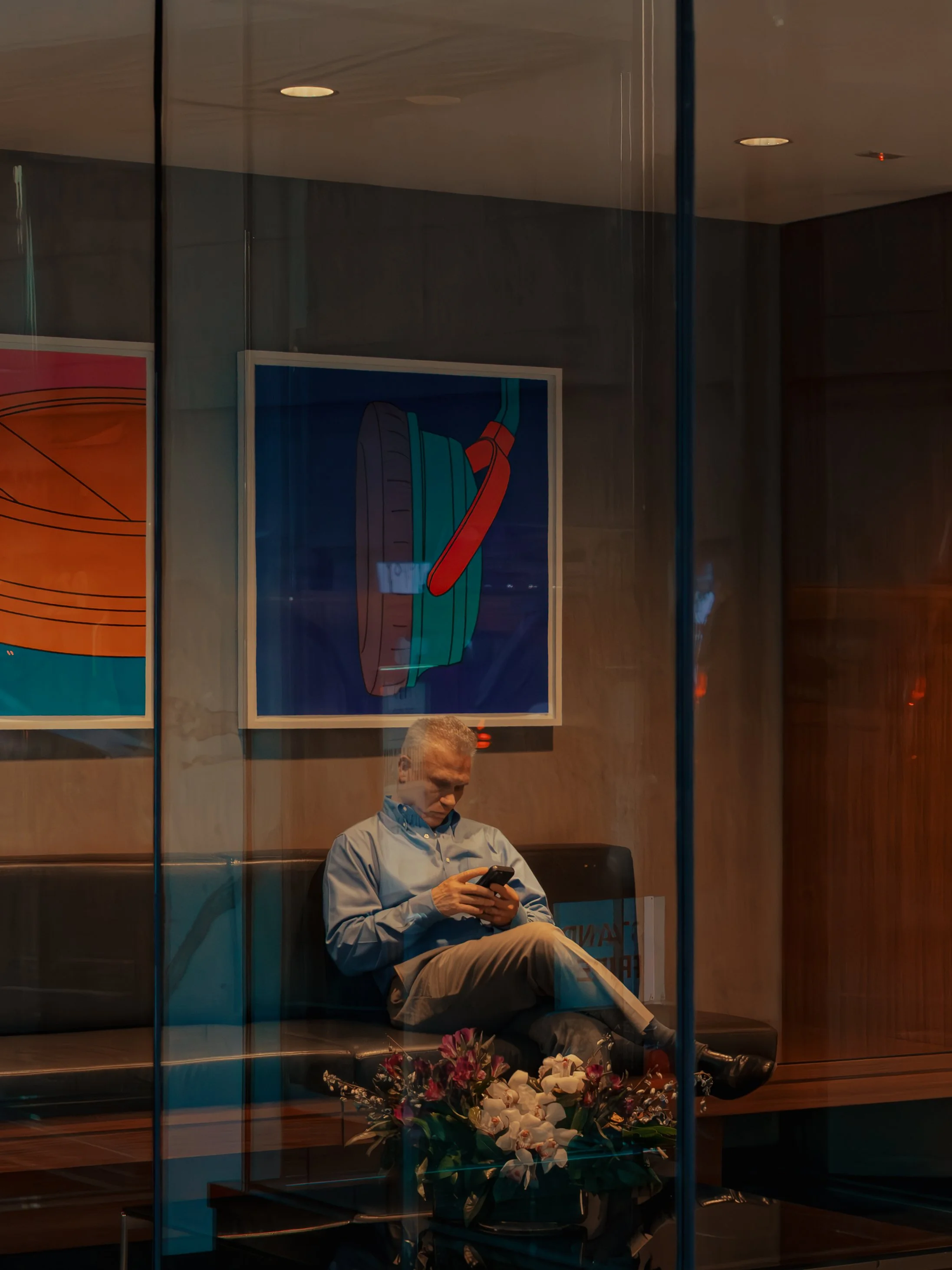 A man sitting in a waiting area, looking at his phone, with floral arrangements in front and colorful abstract art on the wall behind him, seen through a glass window.