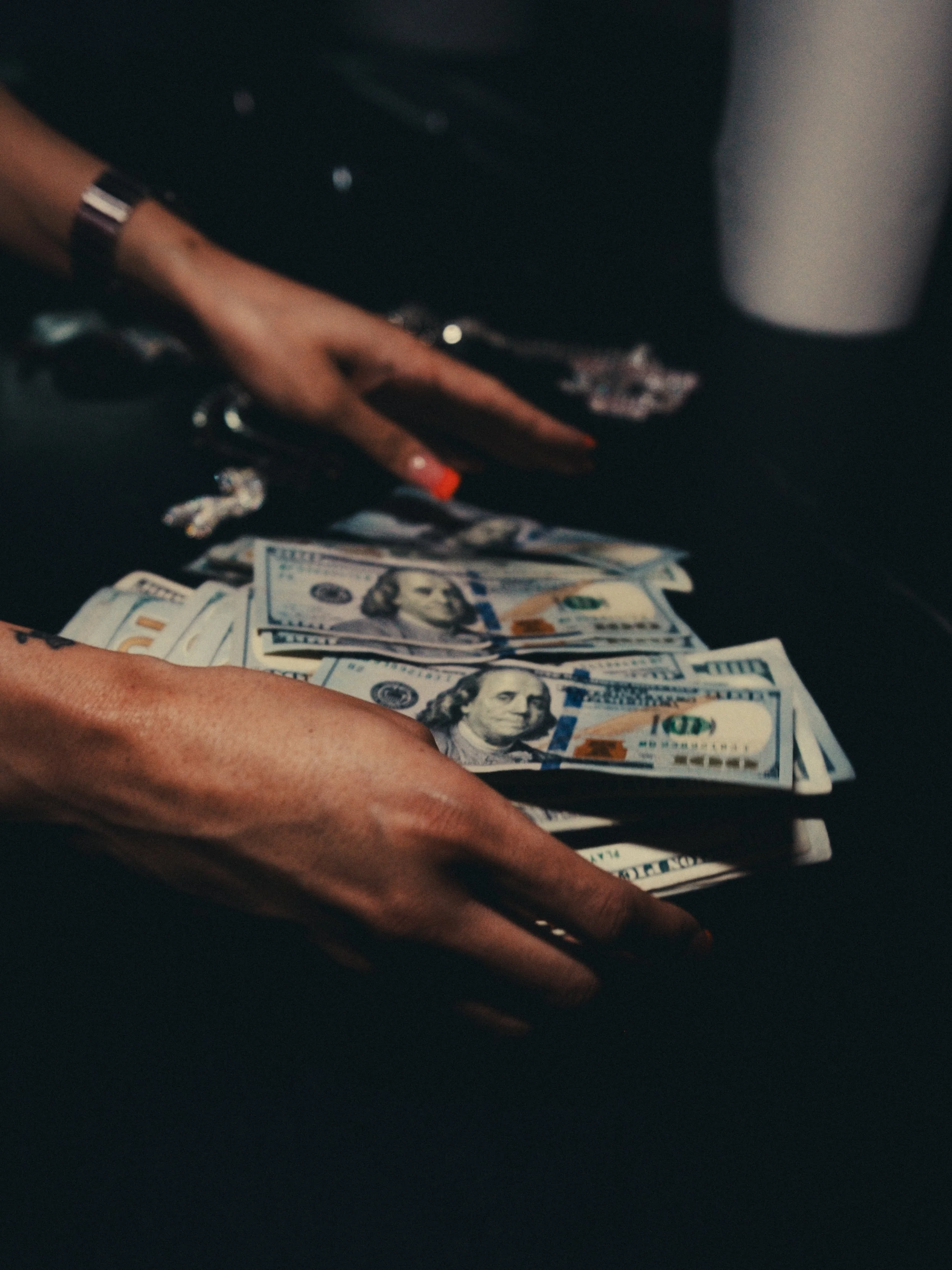 Person's hand with red nail polish counting stacks of hundred-dollar bills on a dark surface.