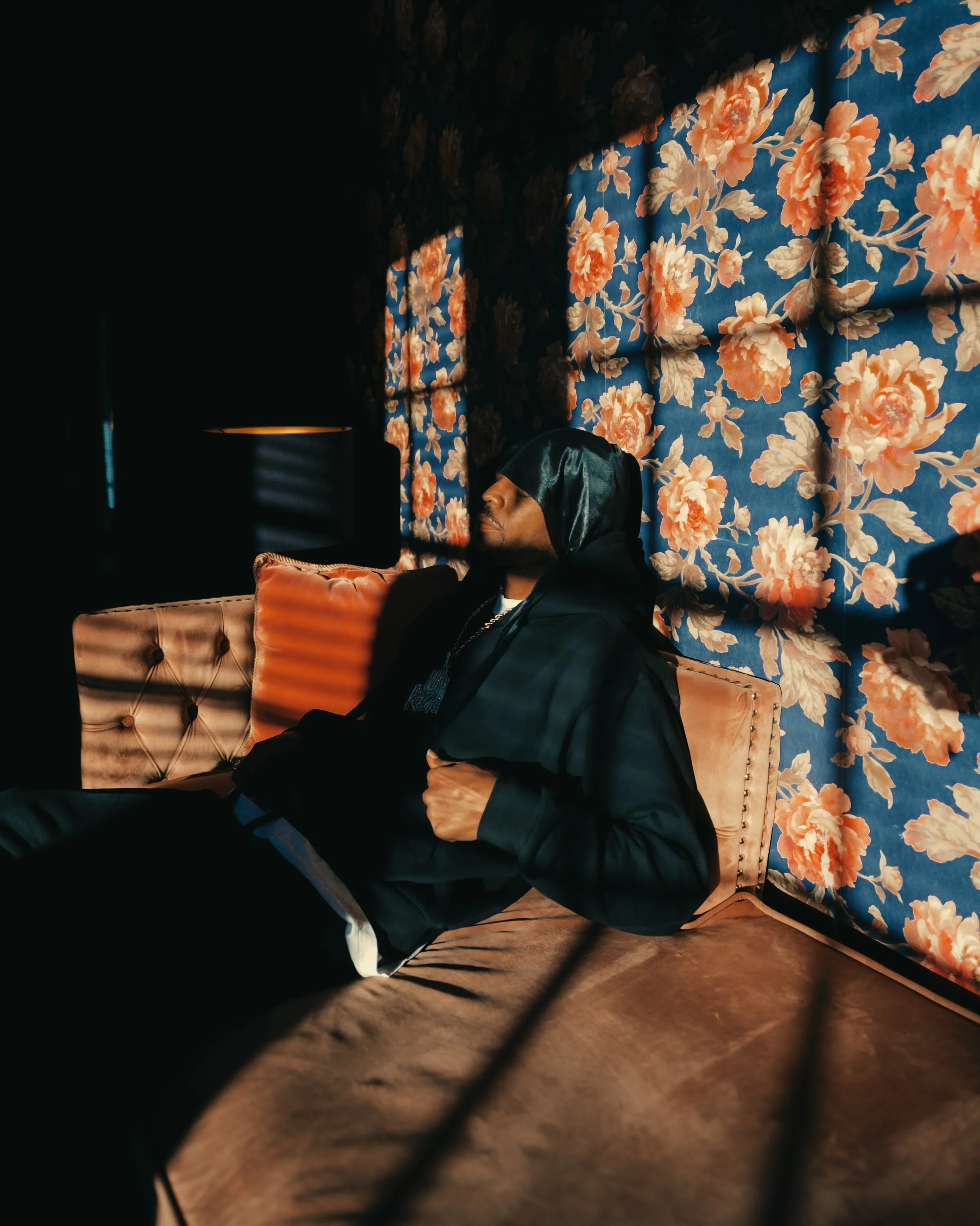A man wearing a black hoodie and head covering, sitting on a tan velvet couch against a floral wallpapered wall. Shadows from window blinds create lines across his face and the wall.