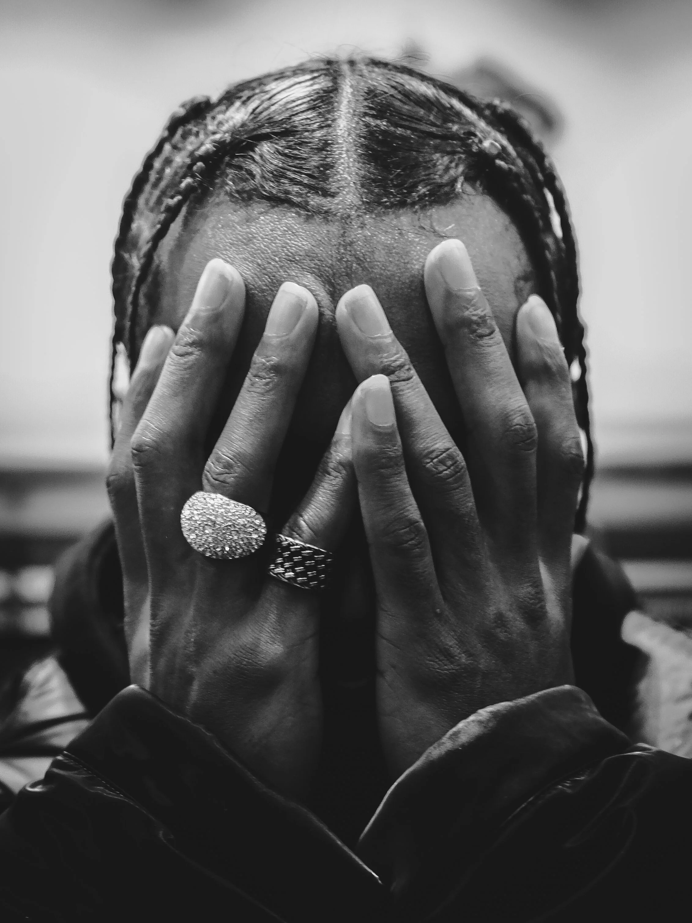 A person with braided hair covering their face with both hands, wearing large rings, and a dark jacket.