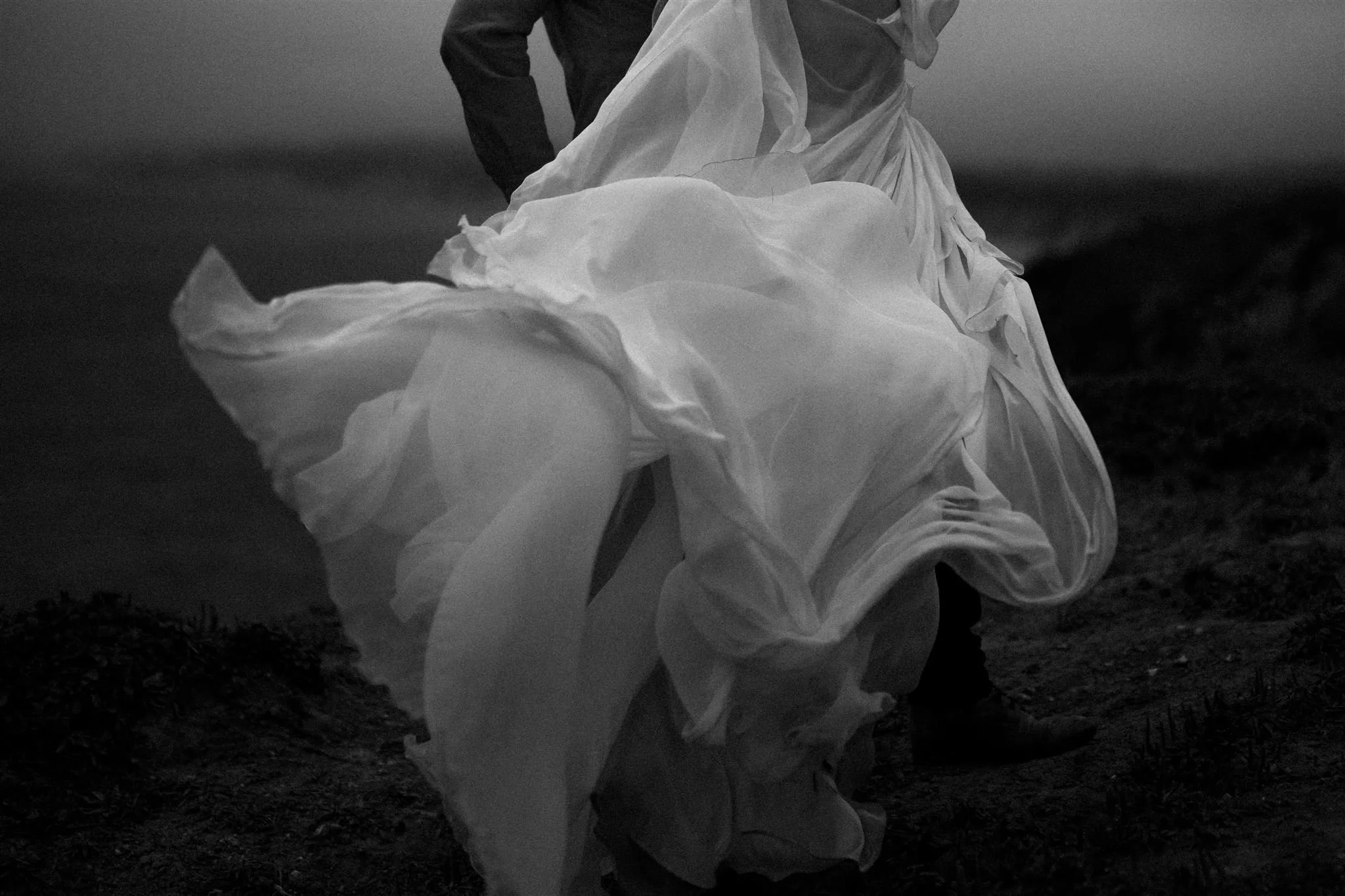 Black and white photograph of a person in a flowing, layered wedding dress walking on a dark, uneven ground in Big Sur, California. Elopement Photography.
