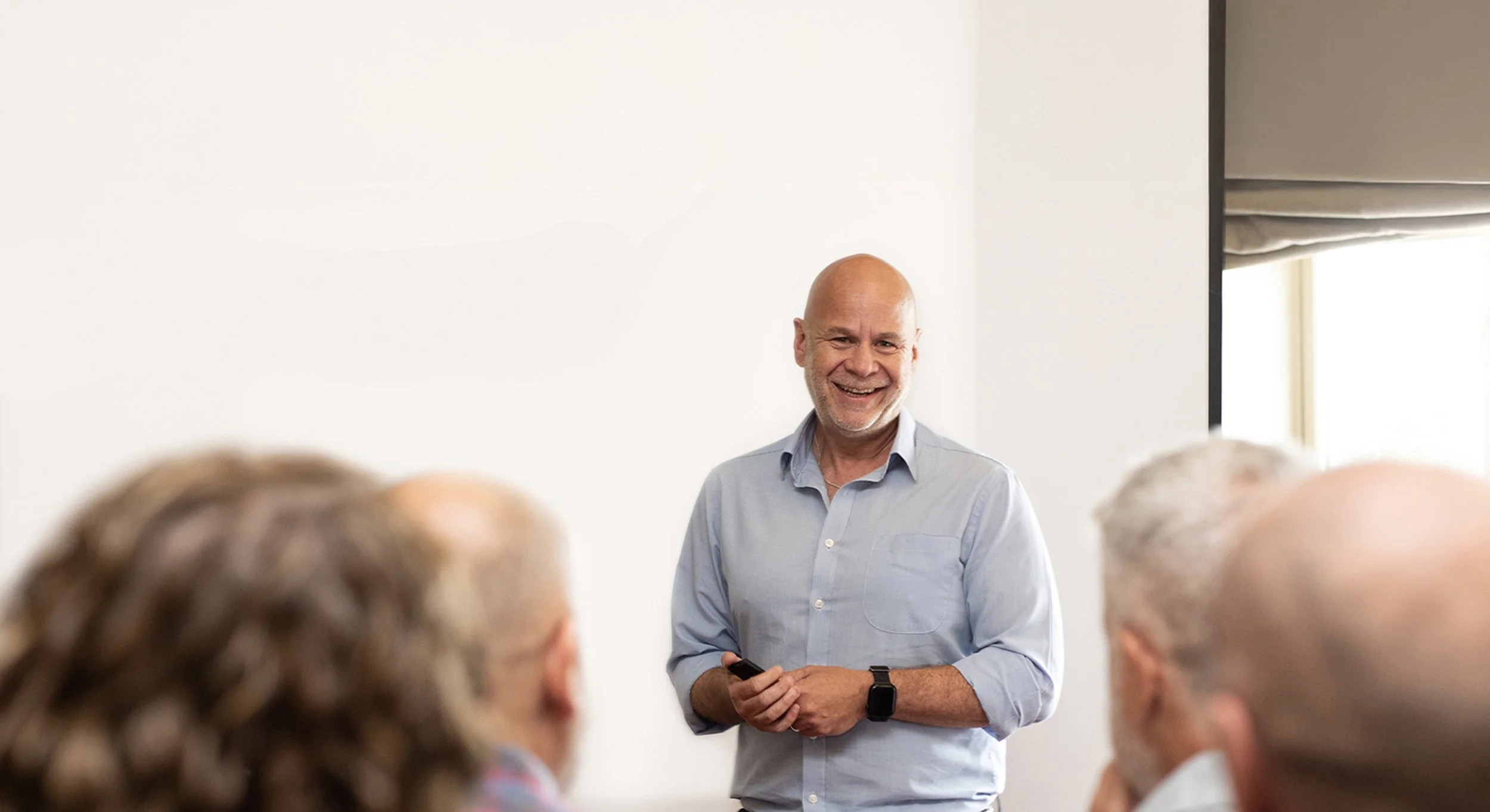 A smiling middle-aged man in a light blue shirt holding a clicker, standing in front of a whiteboard, addressing a group of older adults during a presentation or lecture.