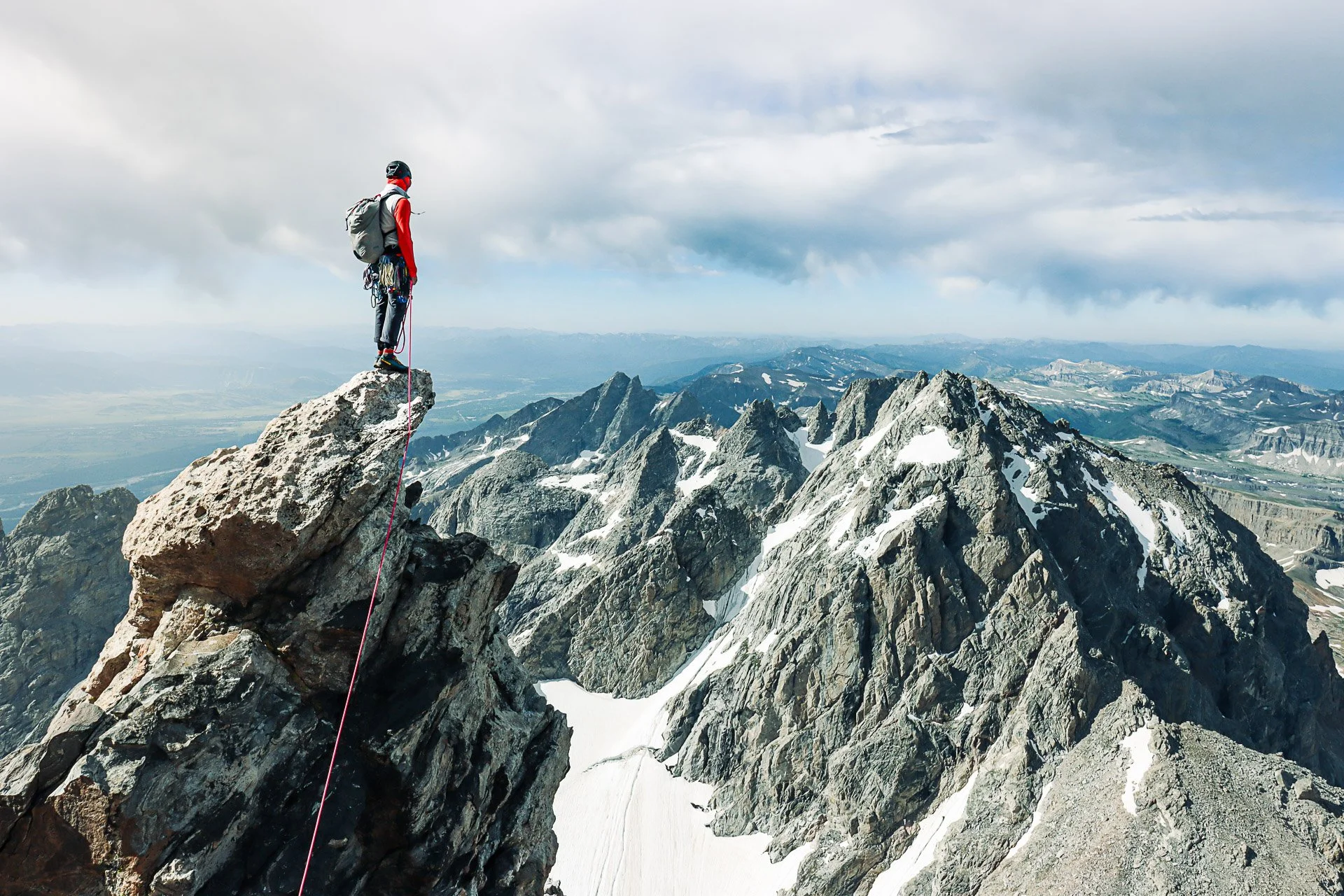 A mountain climber standing on a large rock pinnacle high above snow-capped mountain peaks, with a cloudy sky overhead and a distant valley in the background.