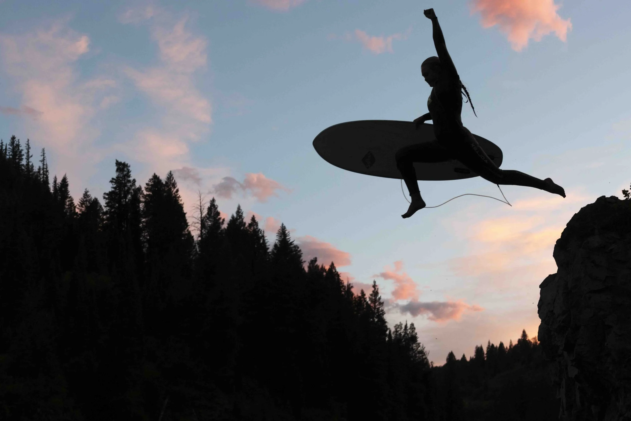 Silhouette of a person mid-air performing a leap with a surfboard, against a sunset sky with pink clouds and a dark forested mountain landscape.