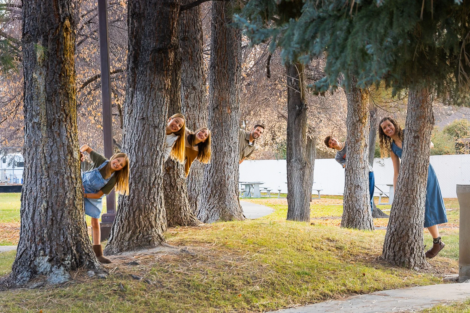 Six young adults peeking from behind trees in a park during autumn.
