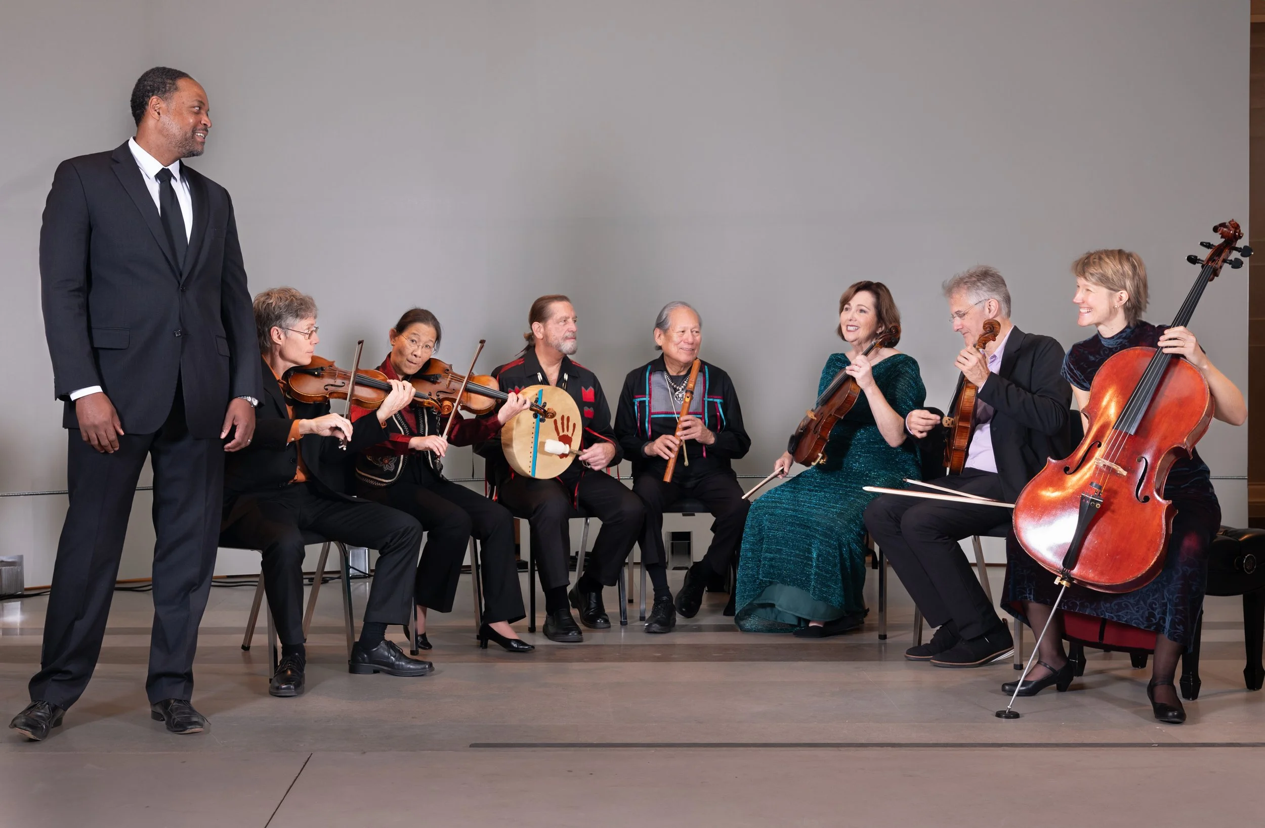 A man in a black suit stands in front of a seated group of diverse musicians holding string instruments, with a neutral gray wall in the background.