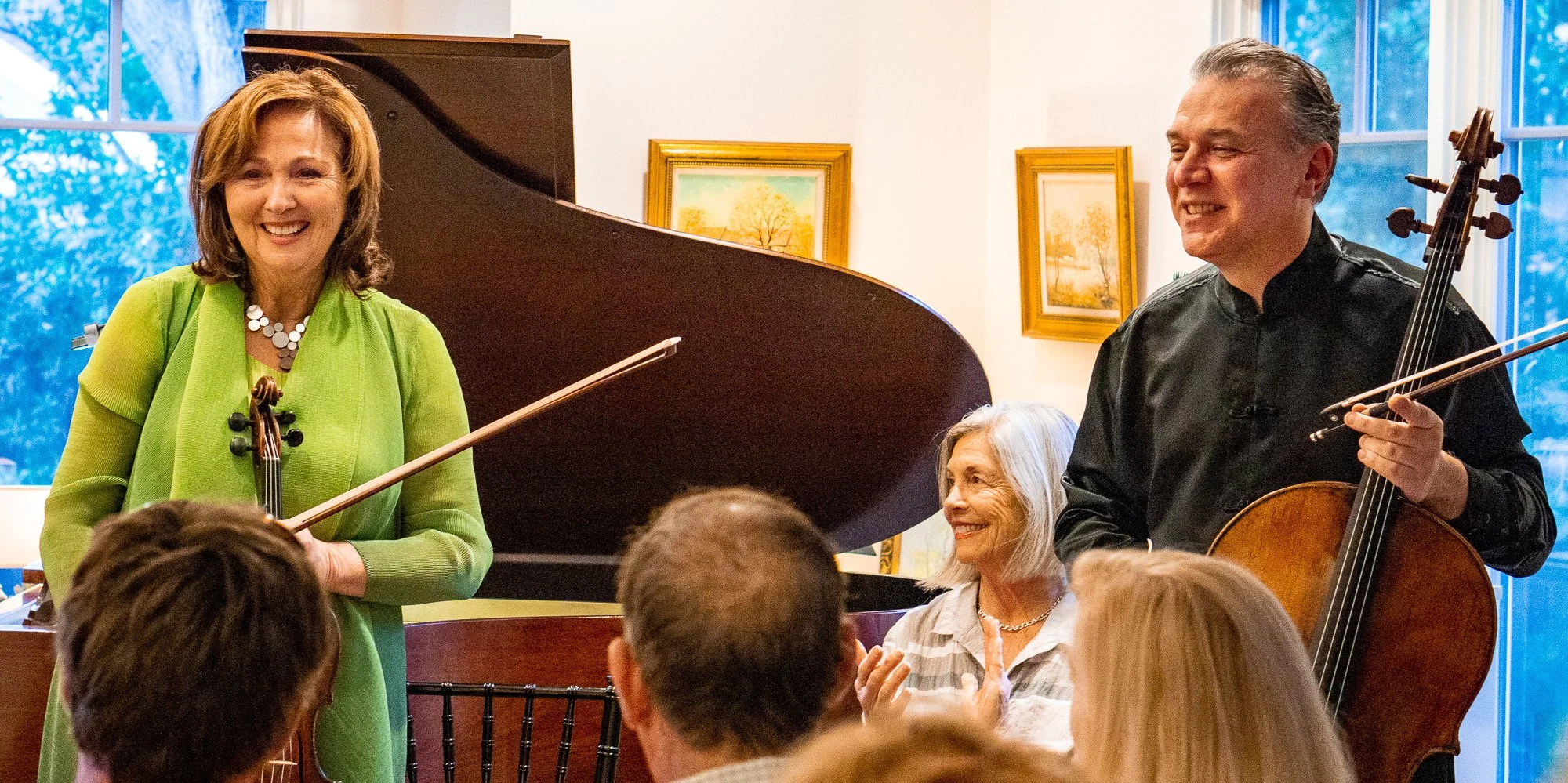 Two musicians, a woman with a violin and a man with a cello, performing in front of an audience. The woman is wearing a green blazer and appears to be smiling, while the man is dressed in black and is also smiling. An audience member, an elderly woman, is clapping and smiling.