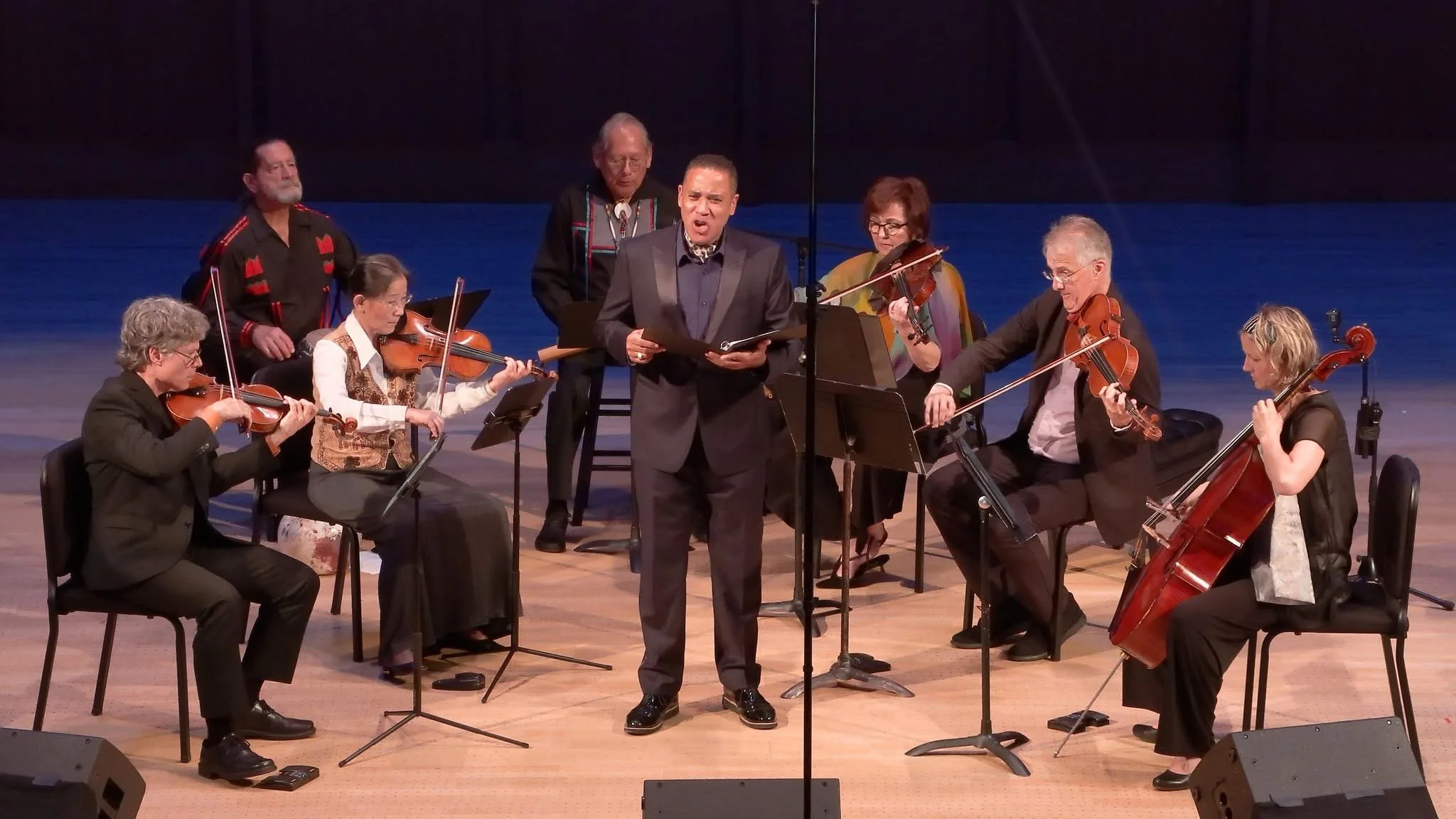 Musicians playing string instruments during a live performance, with a conductor standing in the center and singers in the background.
