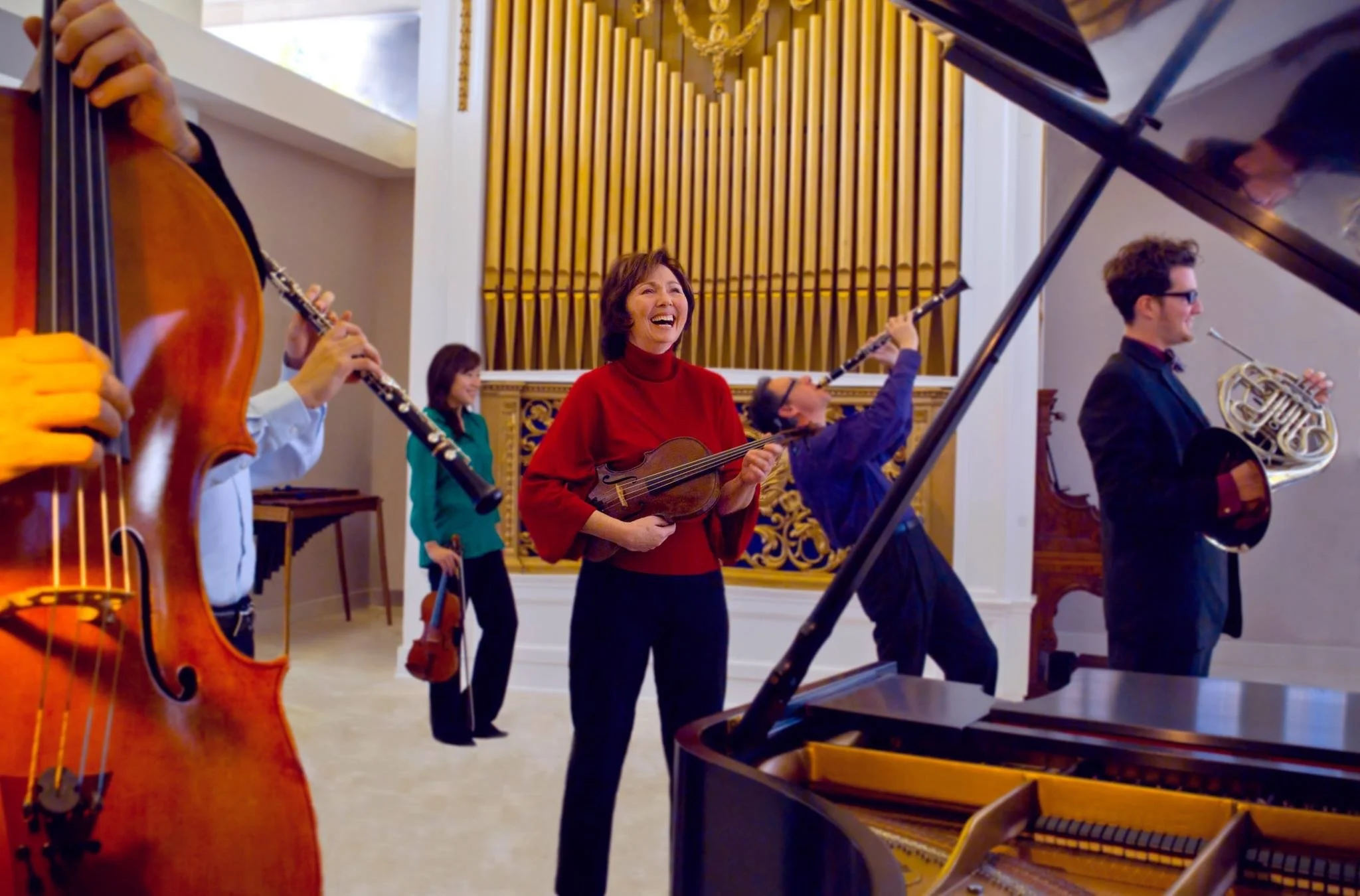 Musicians playing instruments in a bright room with wooden wall accents, including a woman smiling and a grand piano in the foreground.