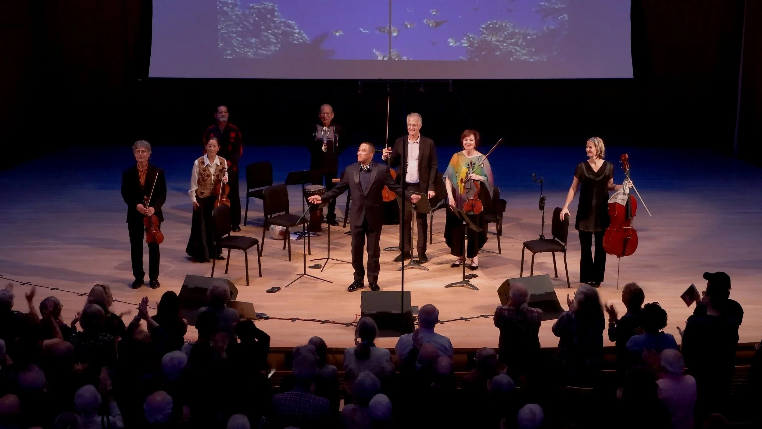 An orchestra on stage with eight musicians, some holding violins and others with a cello, bowing and preparing to play. They are surrounded by chairs, with a large audience in front applauding. A large screen above shows a scenic image of birds flying over trees.