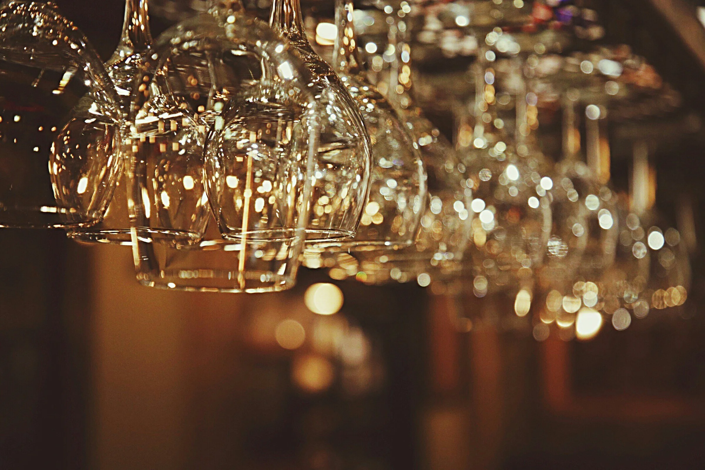 Close-up of upside-down wine glasses hanging upside down in a bar or restaurant setting, with lights creating a bokeh effect in the background.