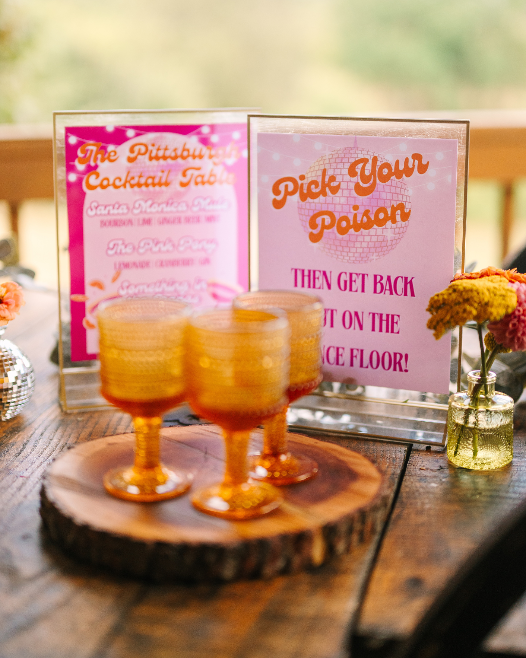 Decorative bar setup with three orange glasses on a wooden slab, pink cocktail menu and a sign reading 'Pick Your Poison' with instructions, and yellow flowers in a small glass jar.