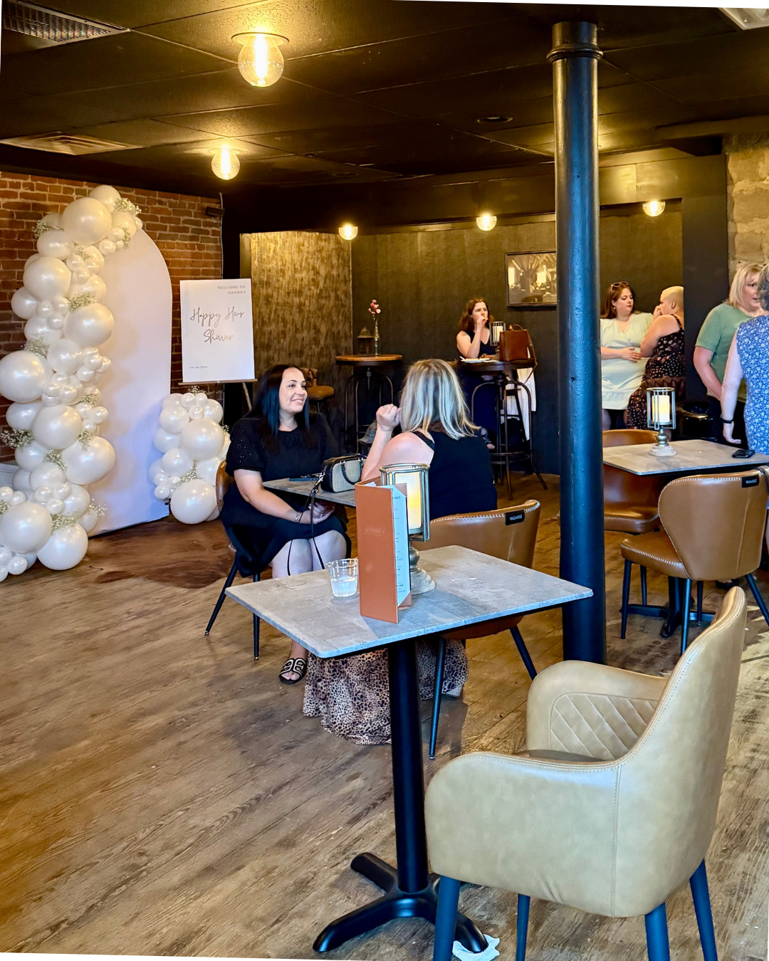 Women socializing and having drinks at a social event in a cozy restaurant or cafe with brick and black walls, decorated with balloon arch and a sign that reads 'Happy Hour Shower'.