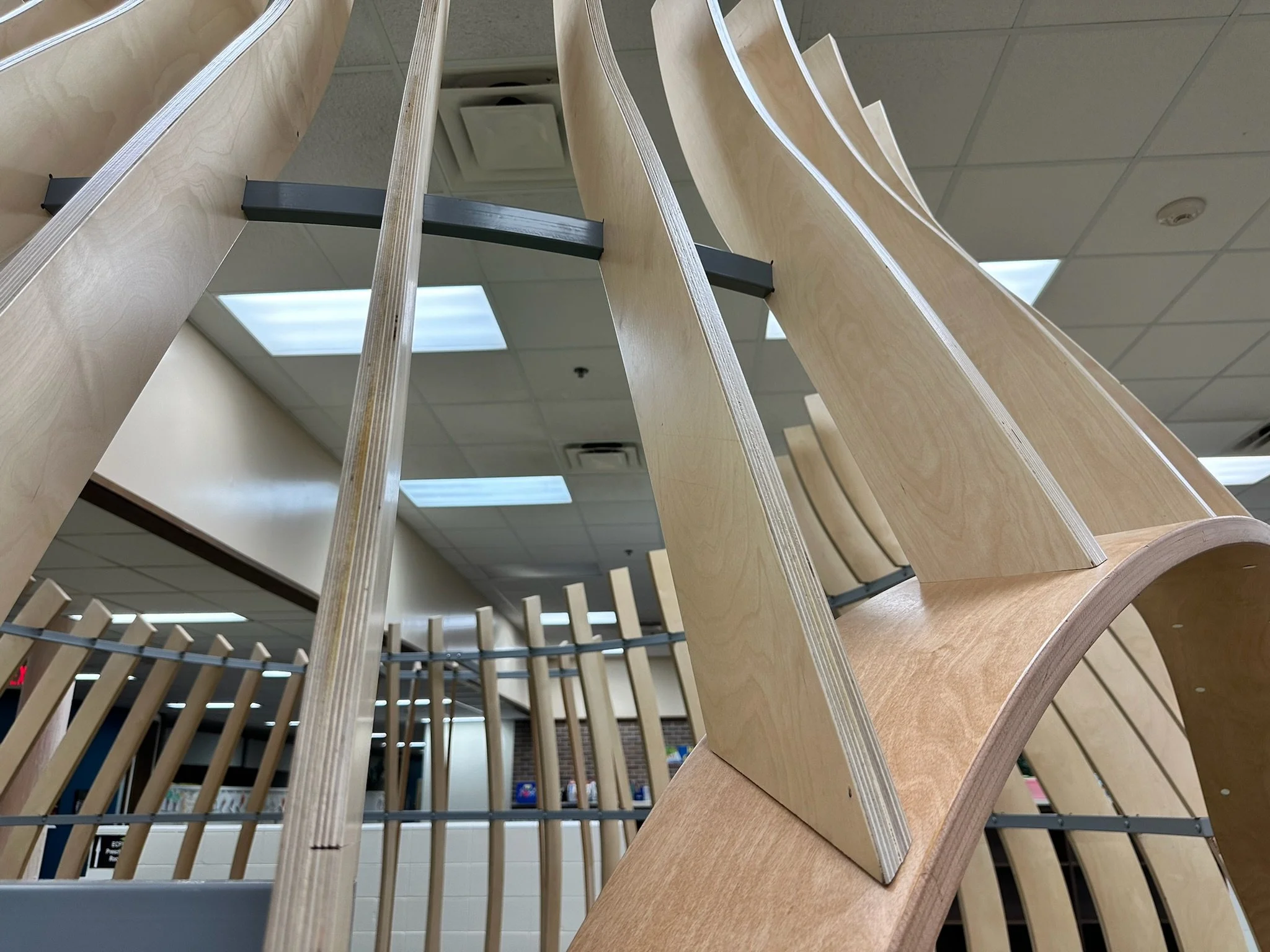 Curved wooden architectural installation with vertical slats indoors, against a ceiling with fluorescent lights.