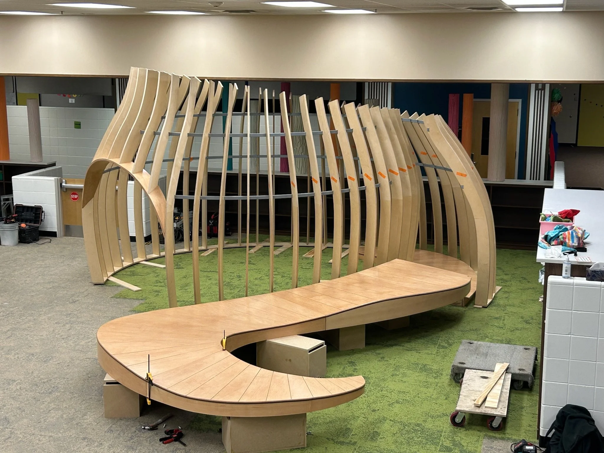 Wooden skeletal structure in a room with green carpet, partially assembled with tools and materials around it.