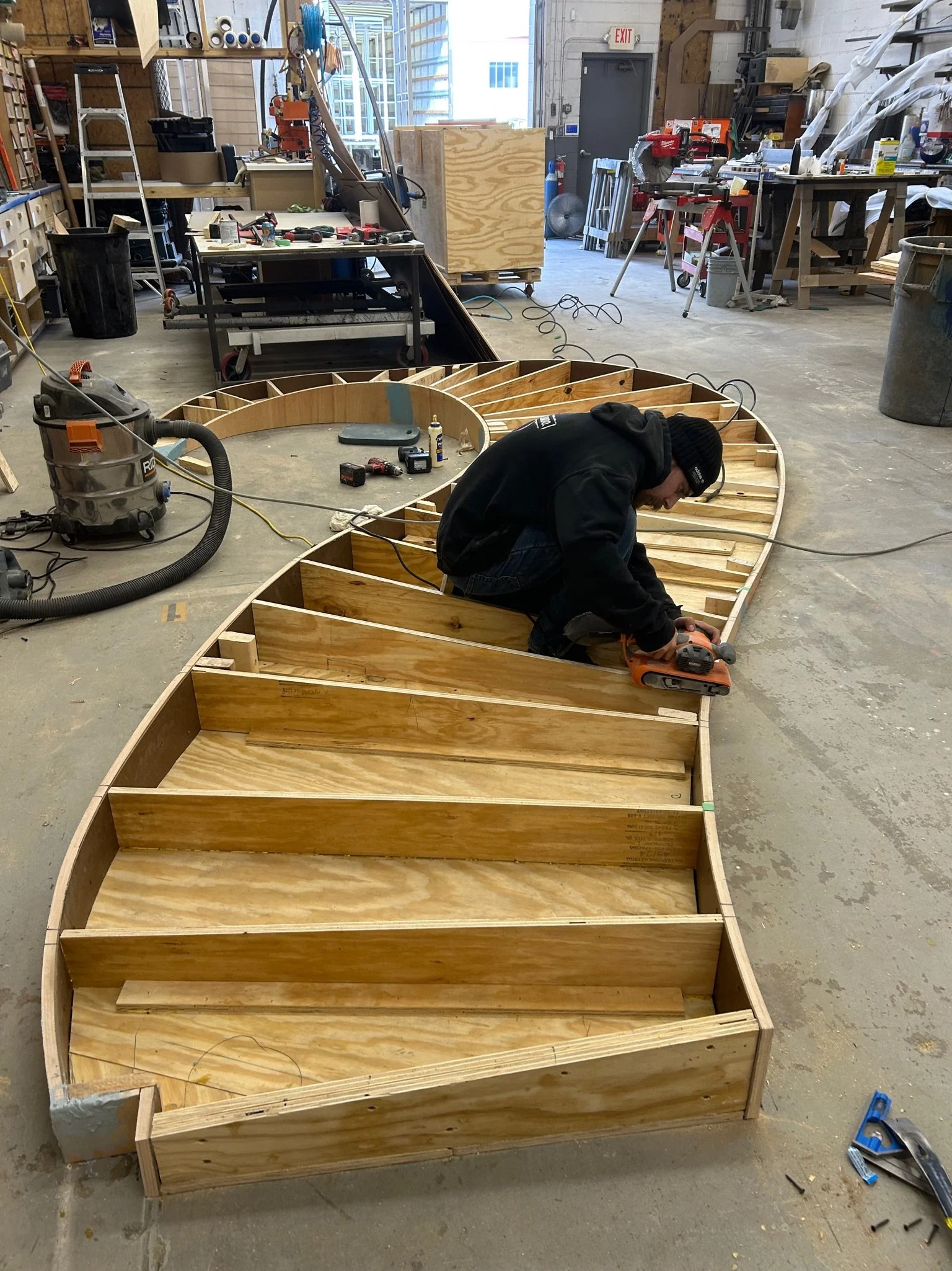 Person working on wooden boat frame in a workshop with tools and equipment.