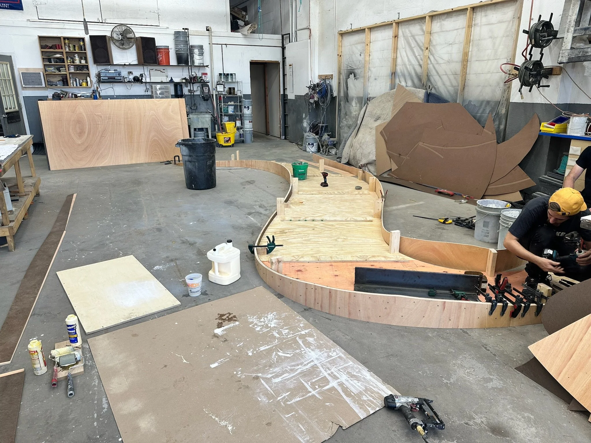 Workshop with partially constructed wooden structure, tools, and materials scattered around. A person in a yellow cap is working on the structure.