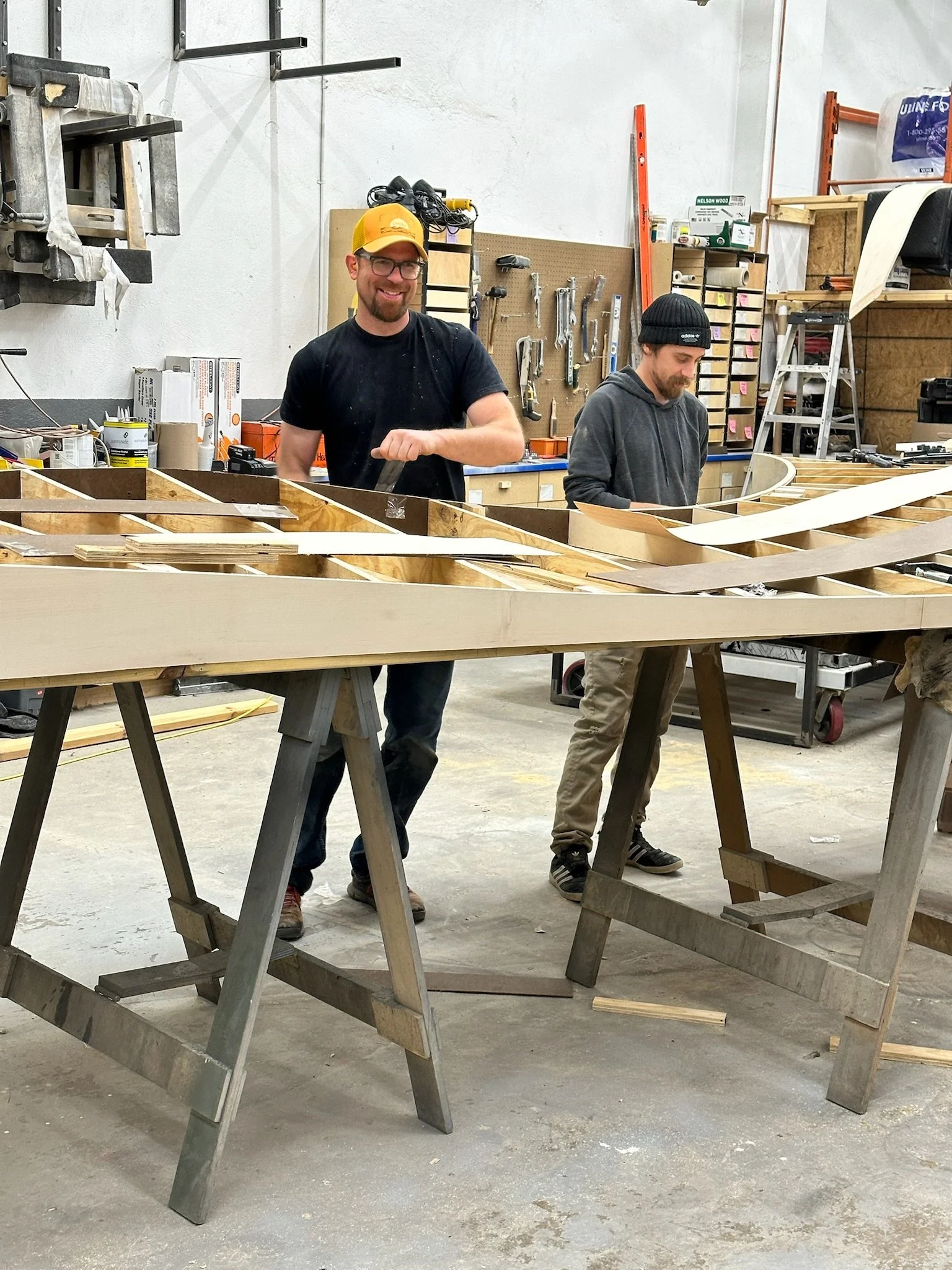 Two people working with wood in a workshop, using sawhorses and tools.