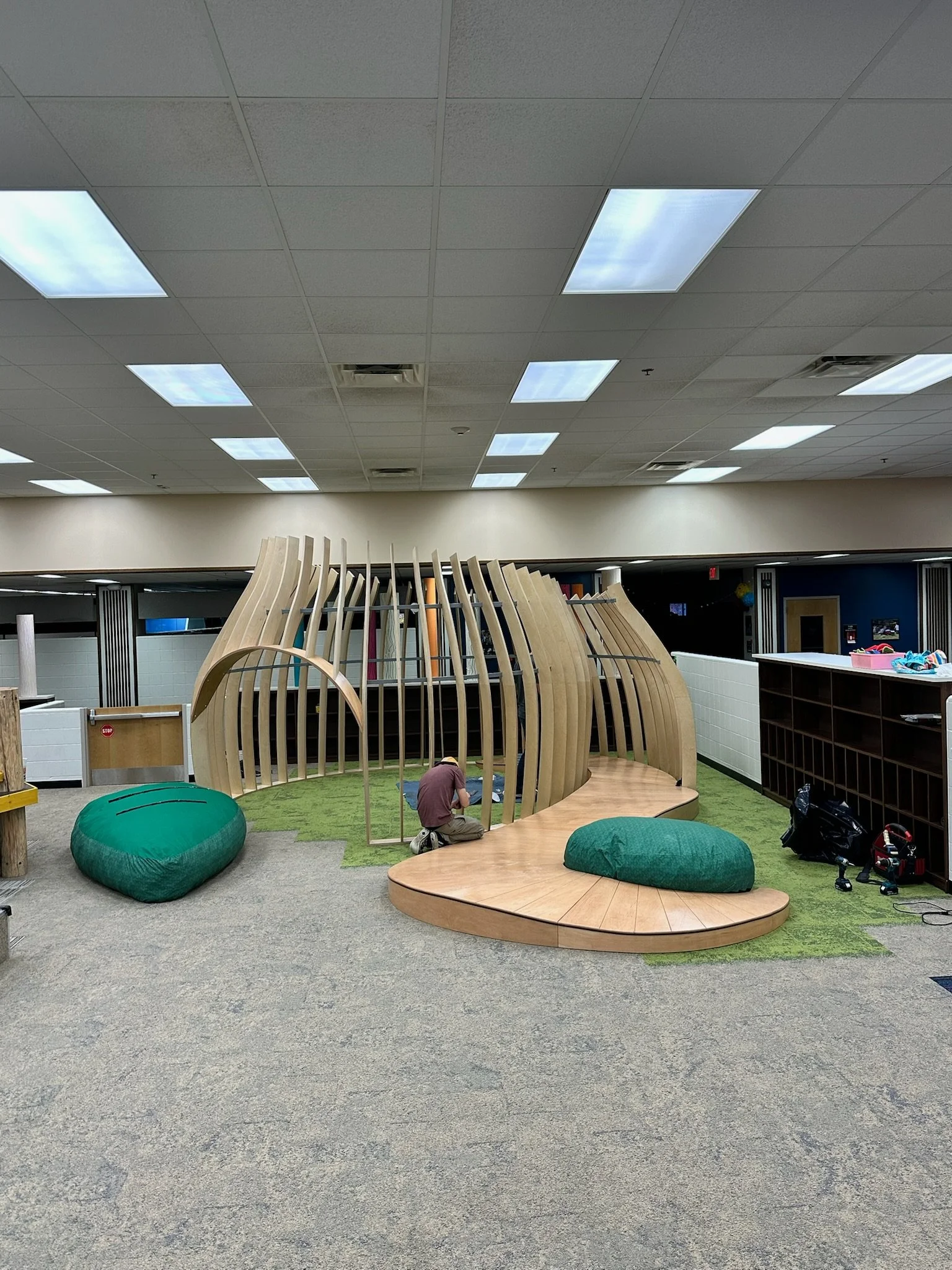 Indoor playground area under construction with wooden play structure, artificial grass, and large green cushions in an open room with a drop ceiling.