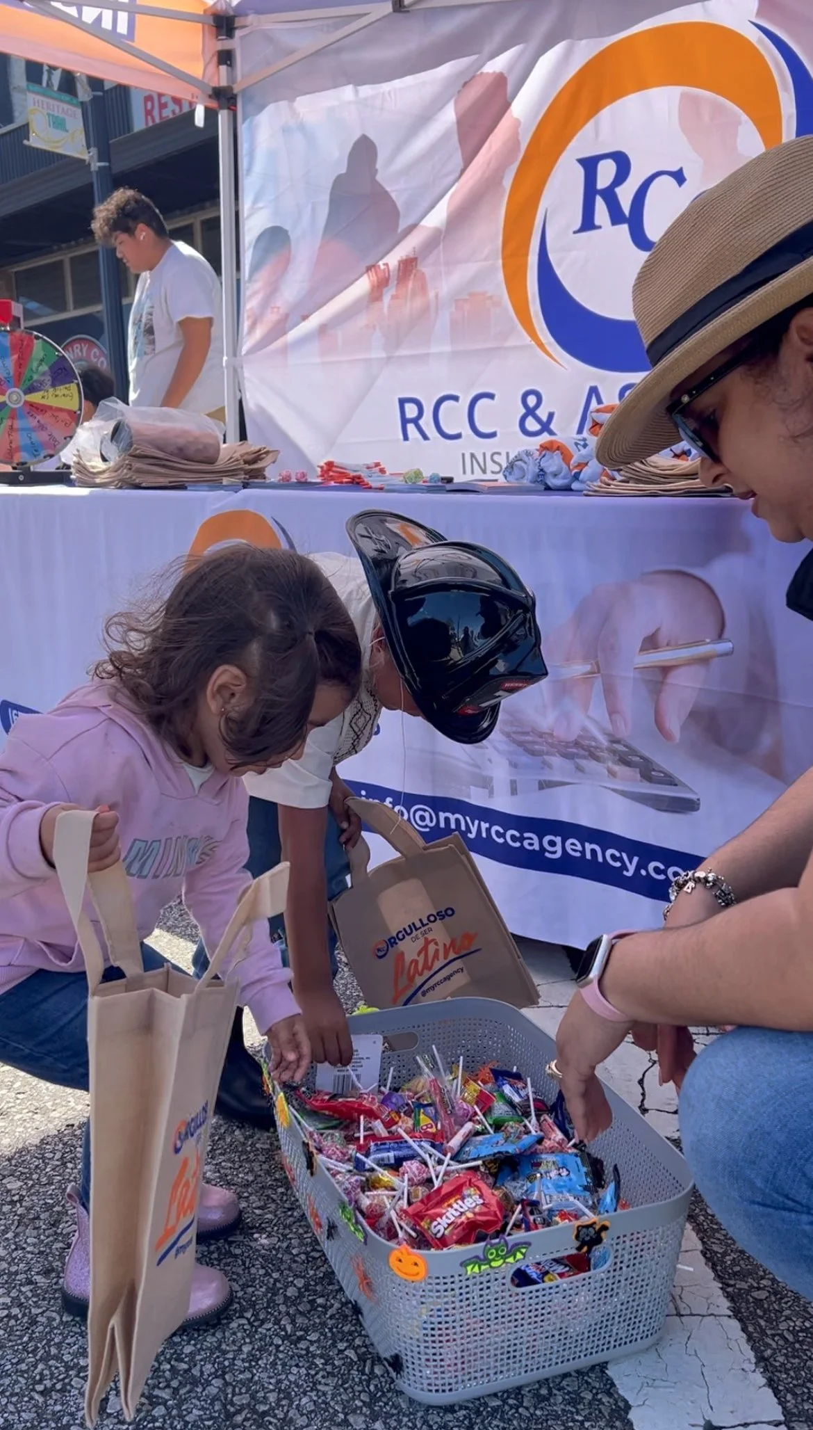 Children selecting candy from a basket at a booth, with adults assisting, under a branded tent featuring promotional materials.