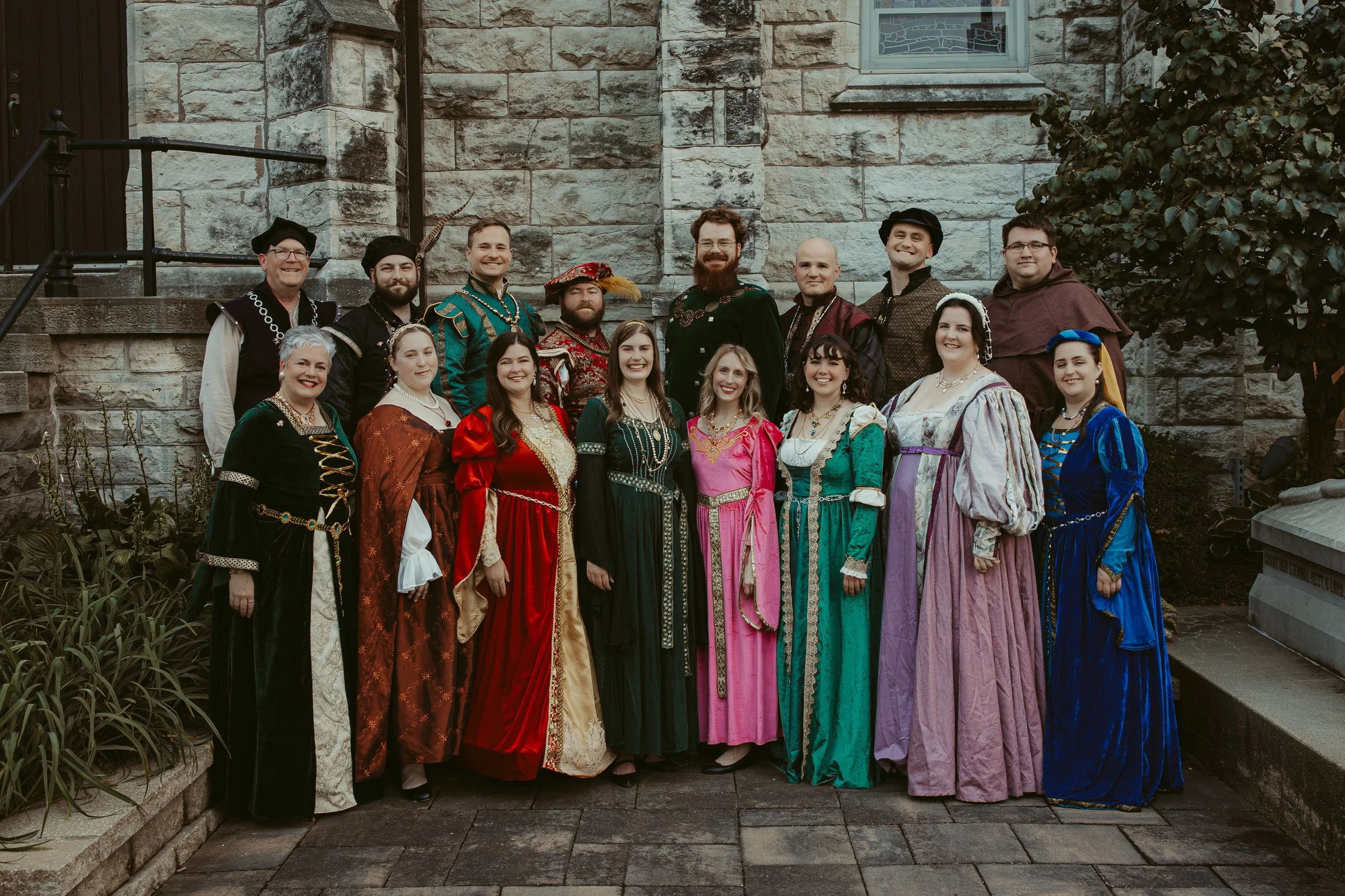 16 people in festive renaissance garb smiling in a cathedral courtyard
