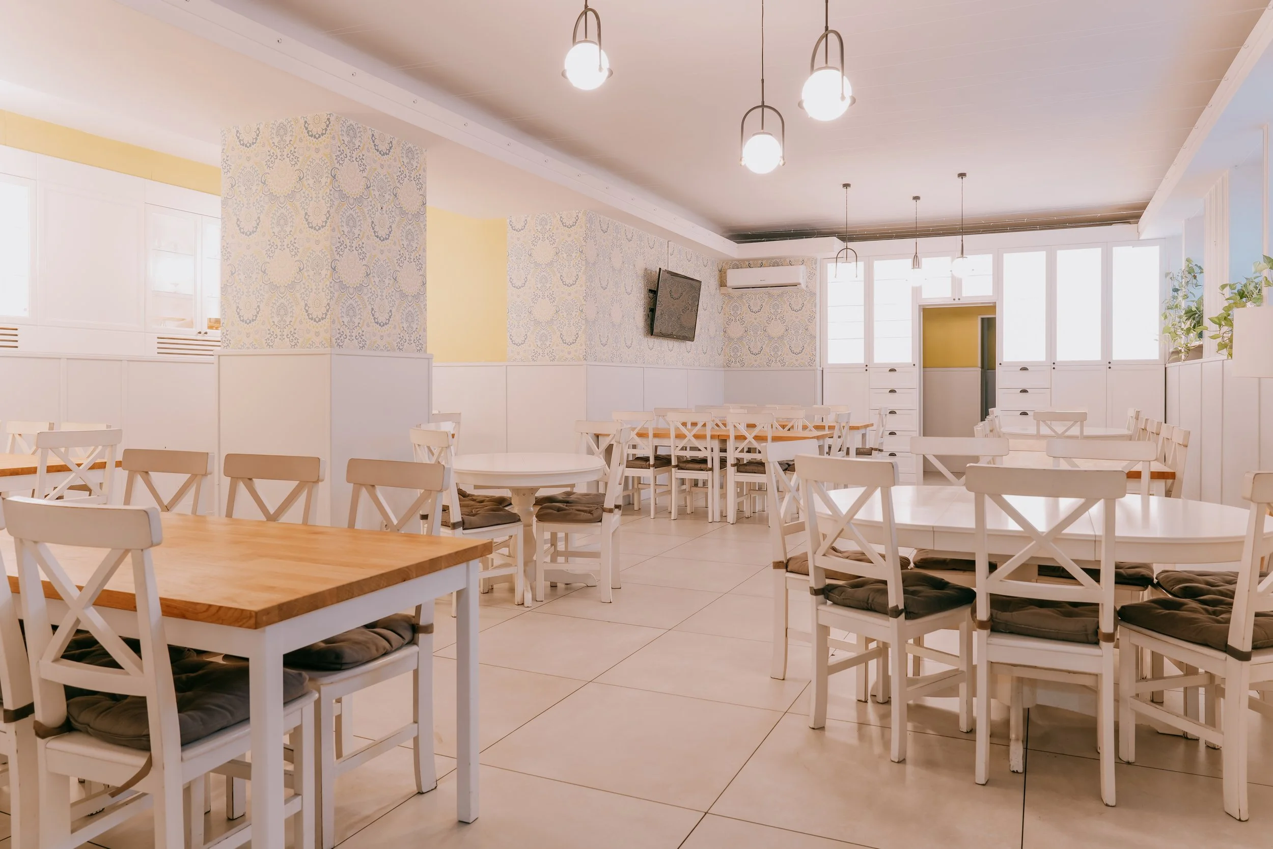 Empty restaurant dining area with white chairs, tables, floral wallpaper, large windows, hanging pendant lights, and an air conditioning unit.