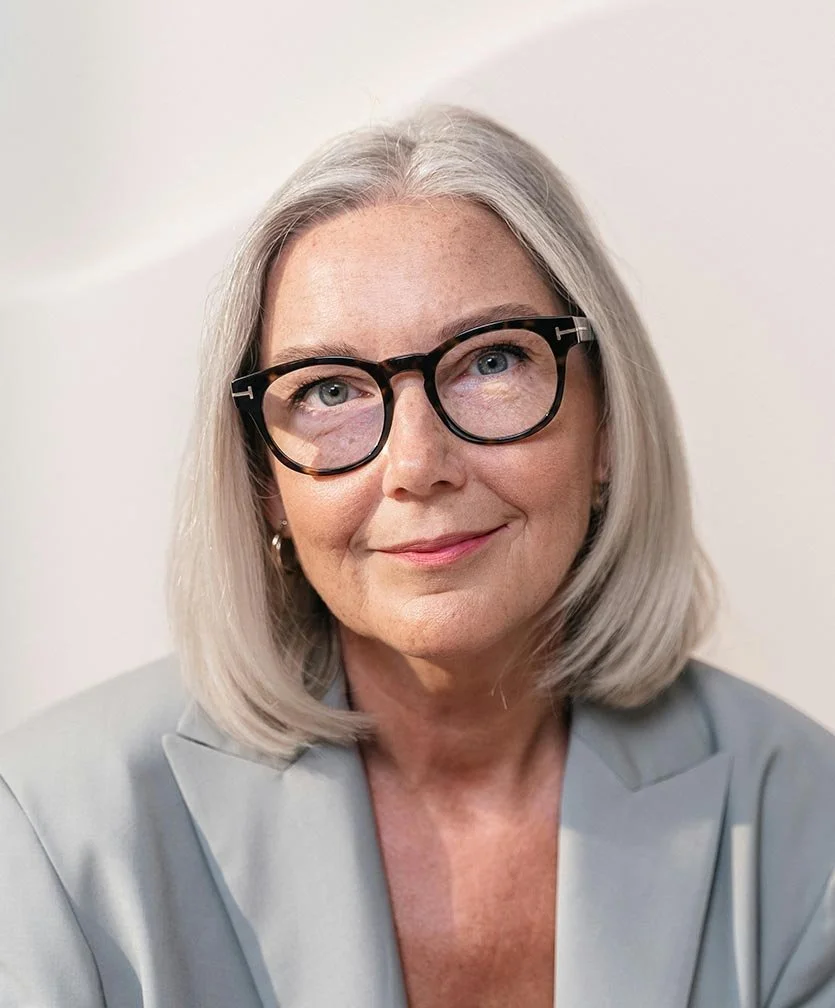 Woman with black glasses and shoulder length gray hair wearing a suit smiles warmly.