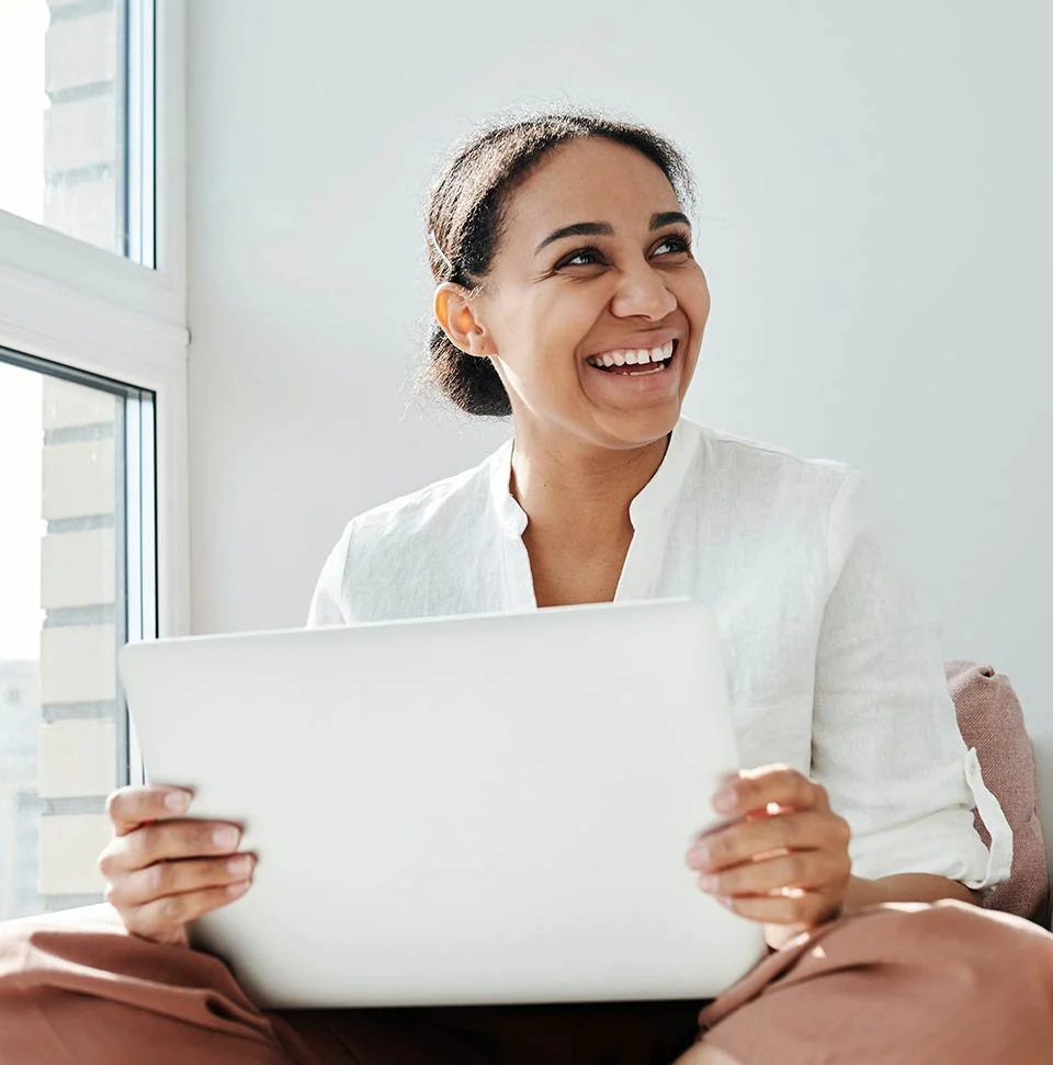Therapist wearing white shirt and peach colored pants sitting near window and holding laptop on her lap while smiling.