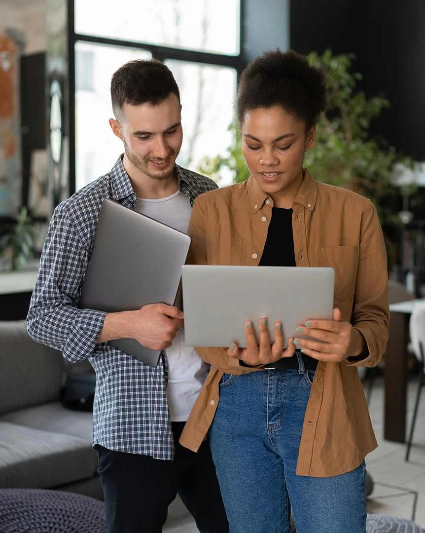 Woman standing holding laptop with a man holding a laptop standing behind her, looking at what's on her computer.