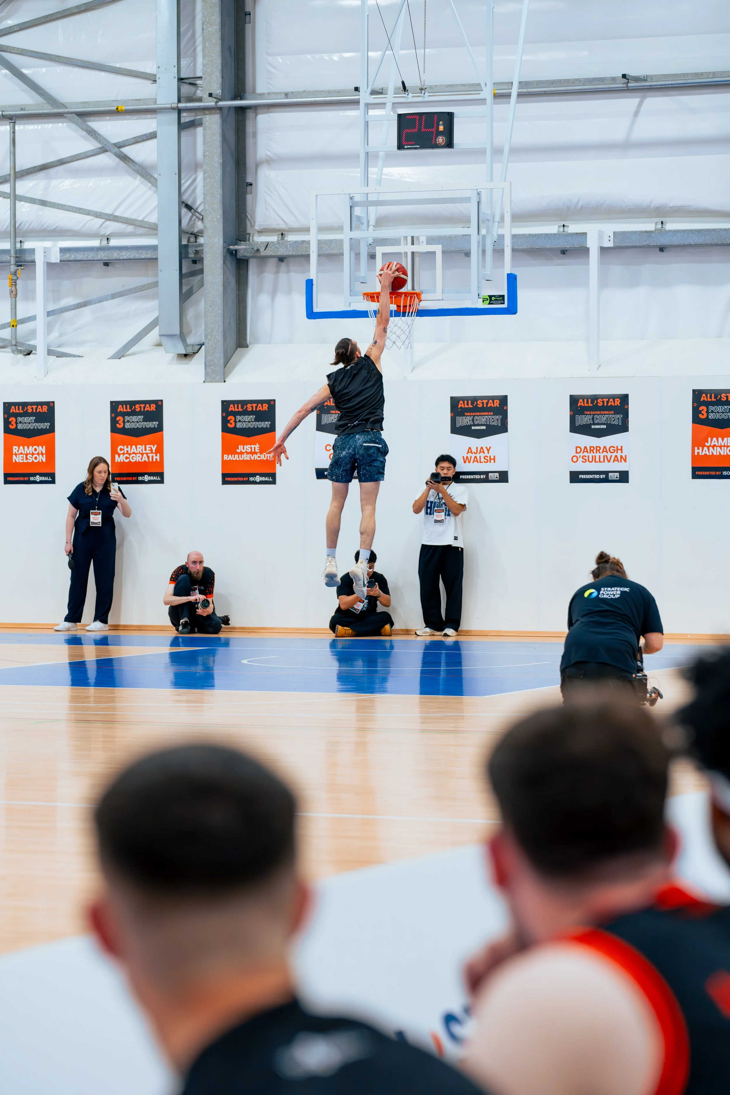 A person in mid-air during a basketball game, about to make a slam dunk on an indoor court. Several people, including photographers and spectators, observe the scene. Courtside Collective Charity Basketball Game 