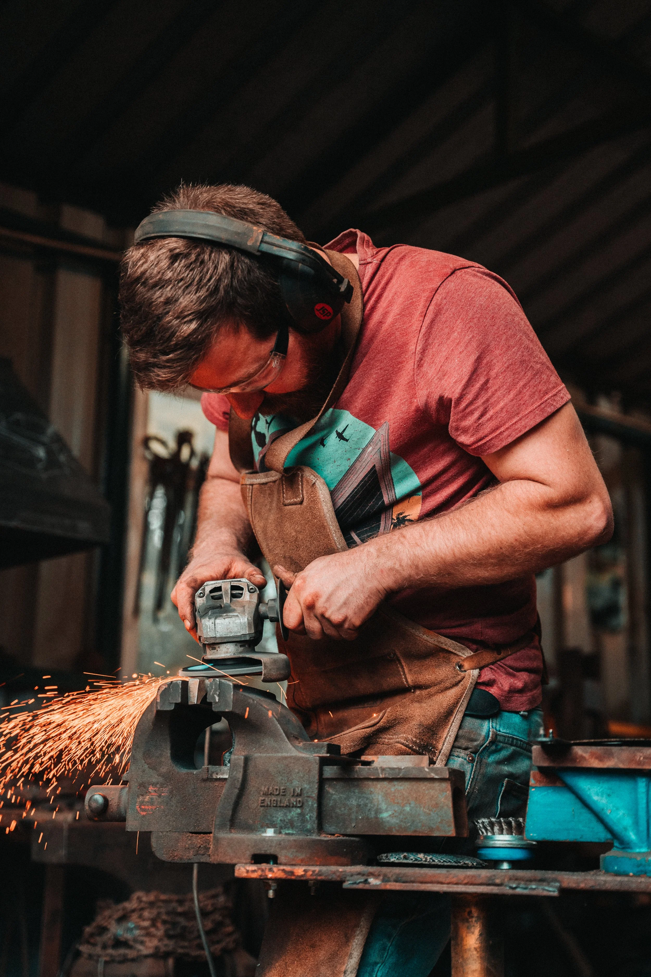 A man welding in a workshop, wearing protective ear muffs and safety glasses, focused on grinding metal with sparks flying.