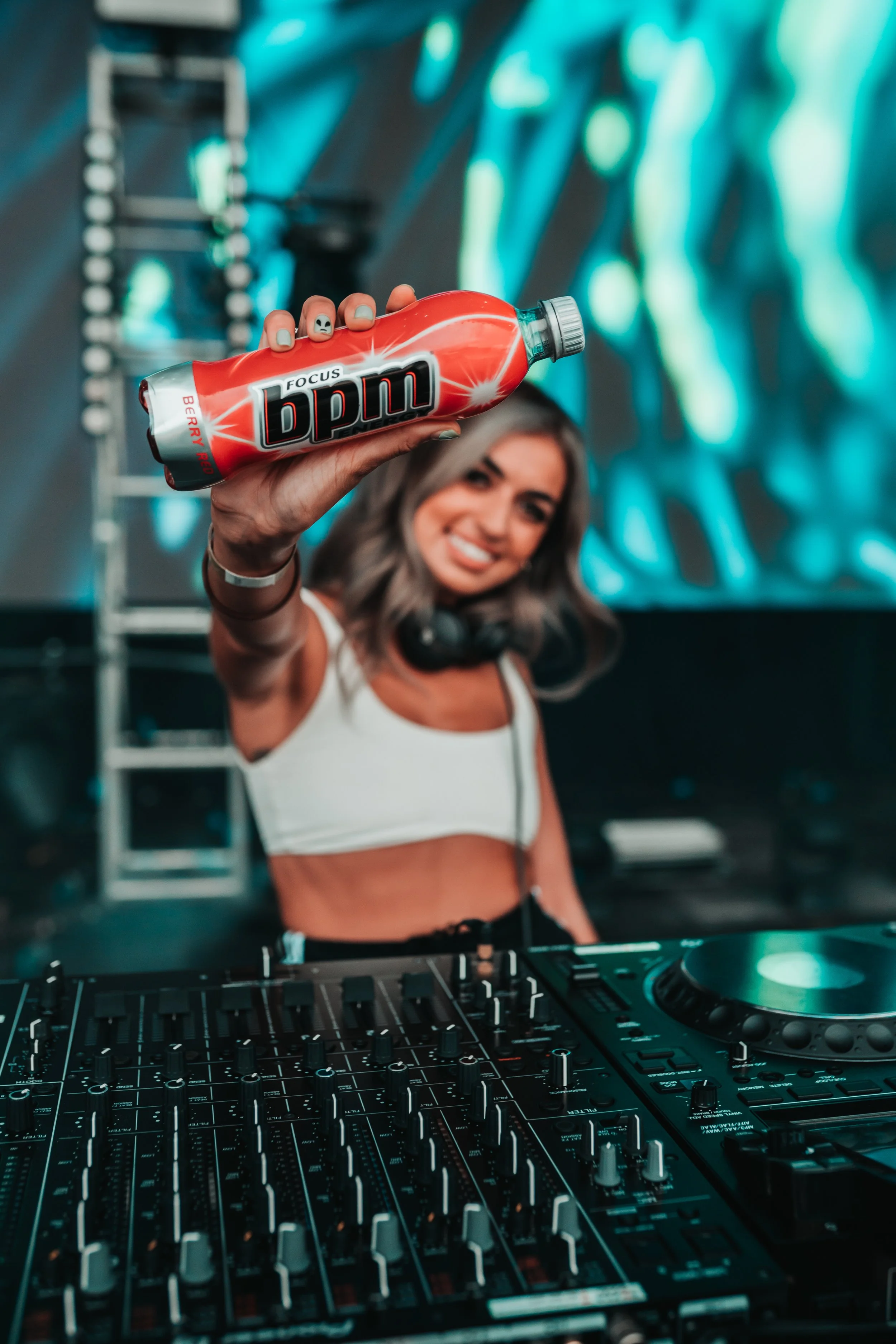 A female DJ with headphones around her neck at a DJ booth, holding a red bottle labeled 'BPM Focus' towards the camera with a smile, in a club or concert environment with colorful background lighting.