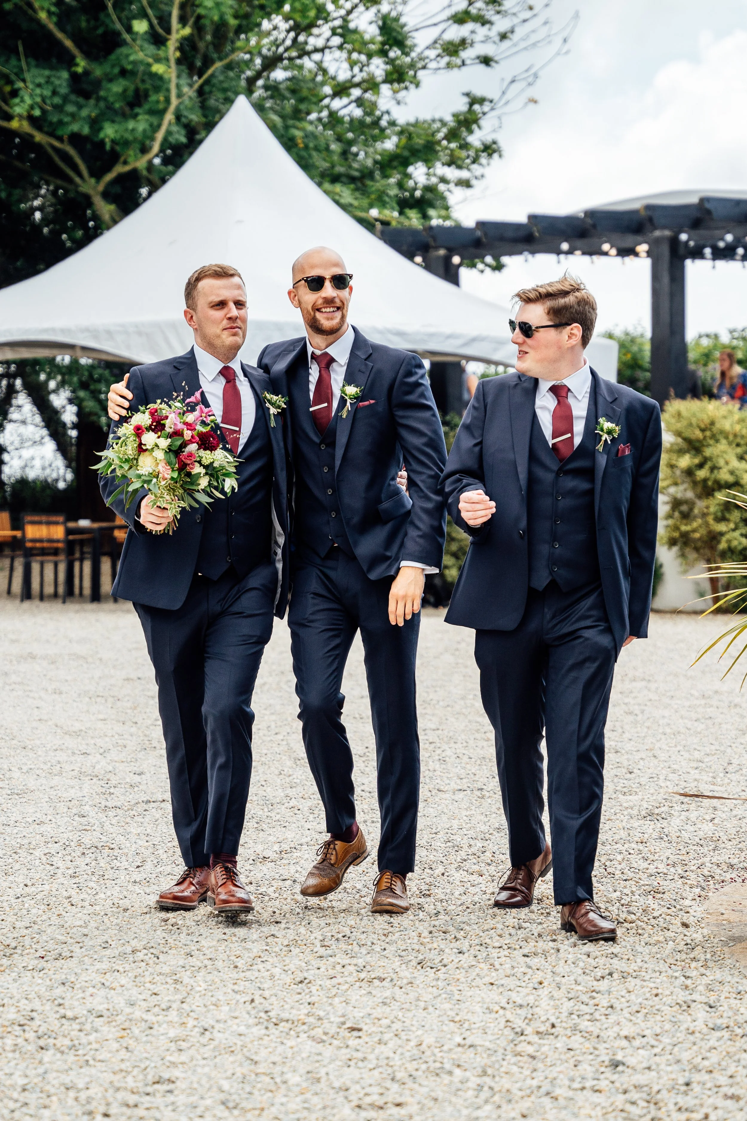 Three men in navy suits with maroon ties walking outdoors at a wedding, with one man holding a bouquet of flowers, smiling, and wearing sunglasses.