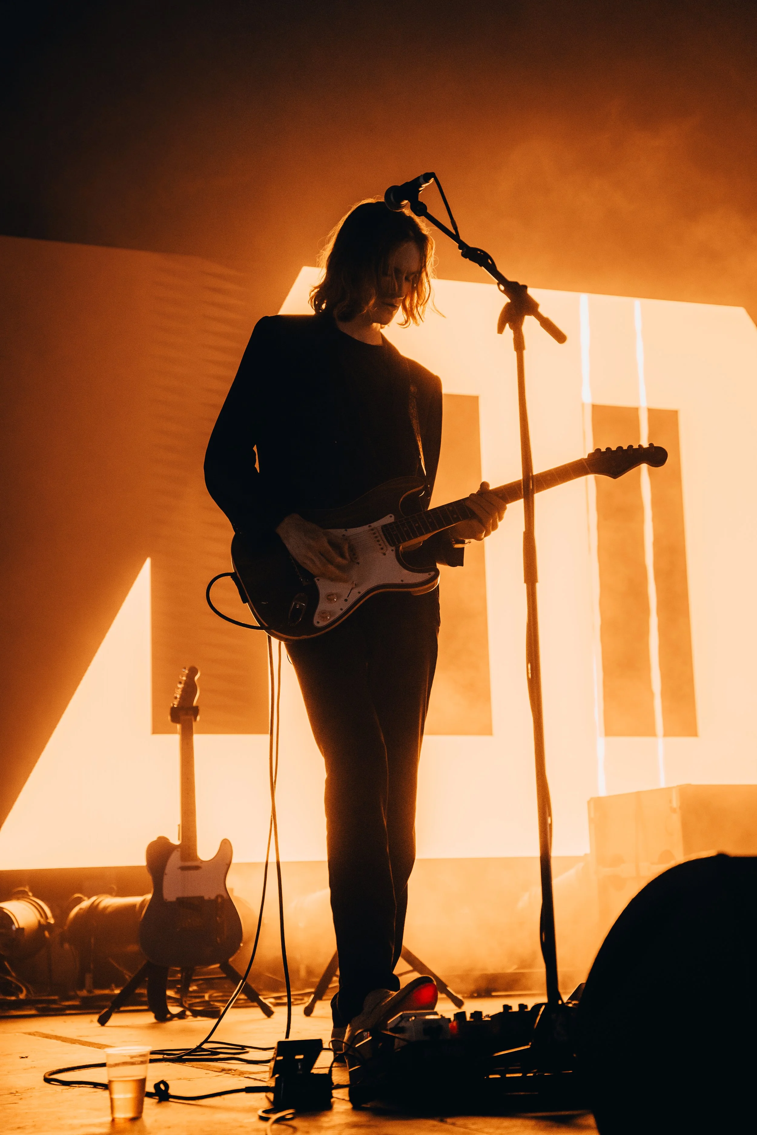 A musician playing an electric guitar on stage with orange lighting and a large illuminated backdrop behind them.