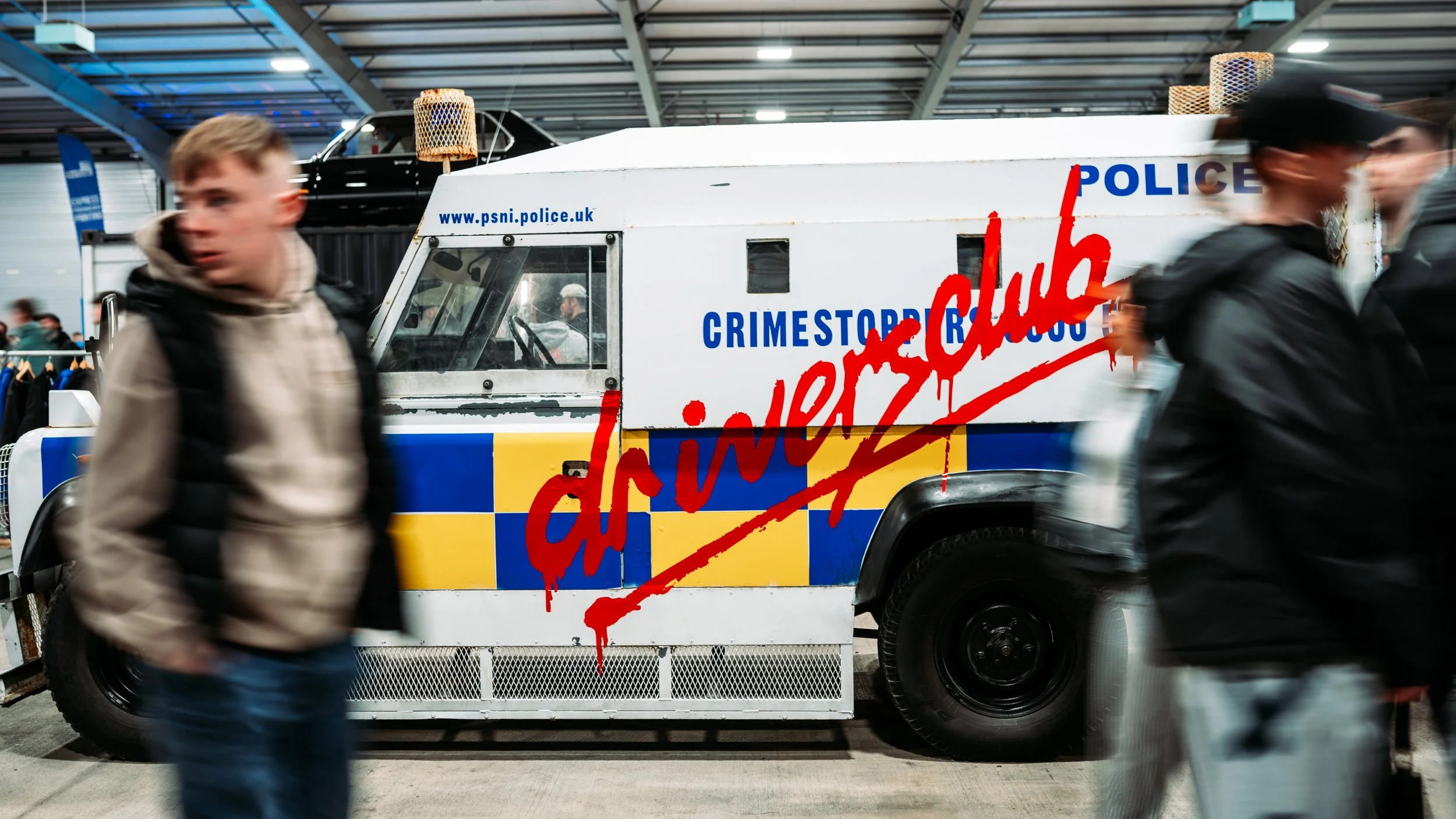 People walking past a police vehicle with the words 'CRIMESTOPPERS' and 'challenges' written in red graffiti-style text on the side, at an indoor event or exhibition.