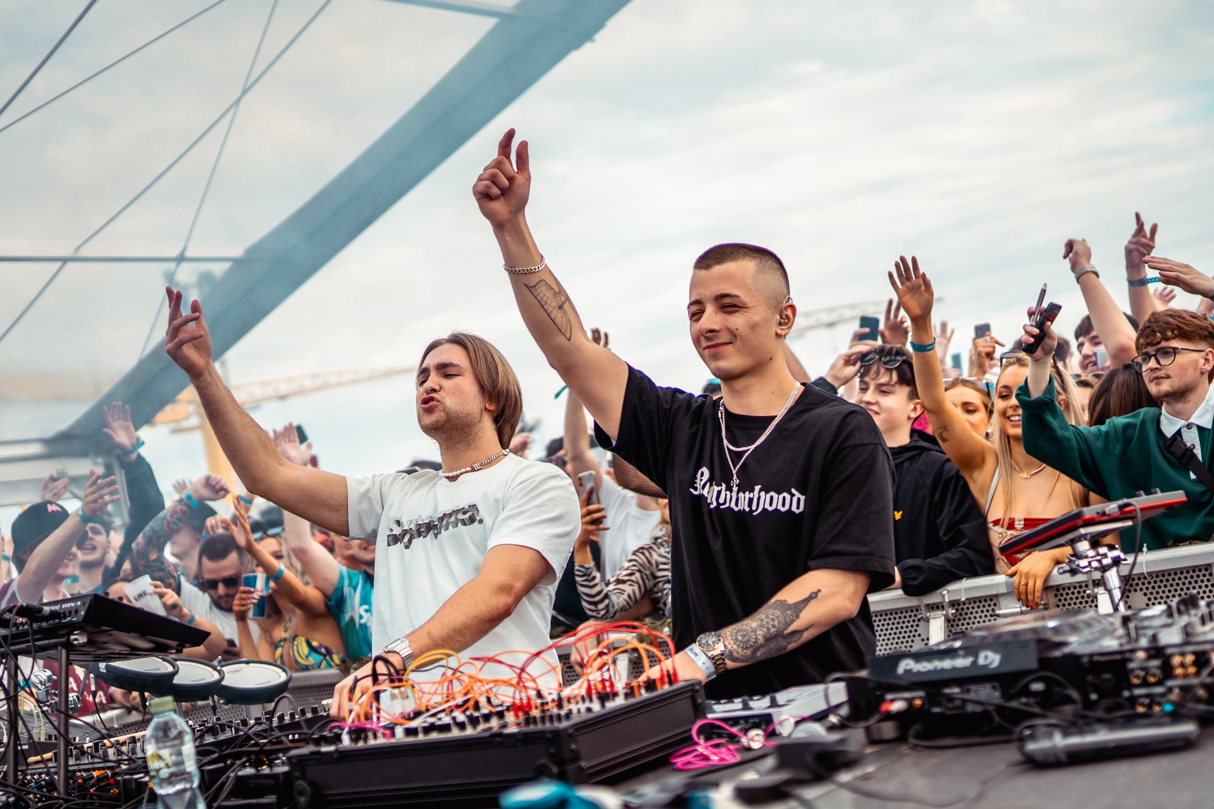 Two DJs perform on an outdoor stage at a music festival, surrounded by a crowd of cheering fans, with some raising their hands and phones, under a partly cloudy sky.