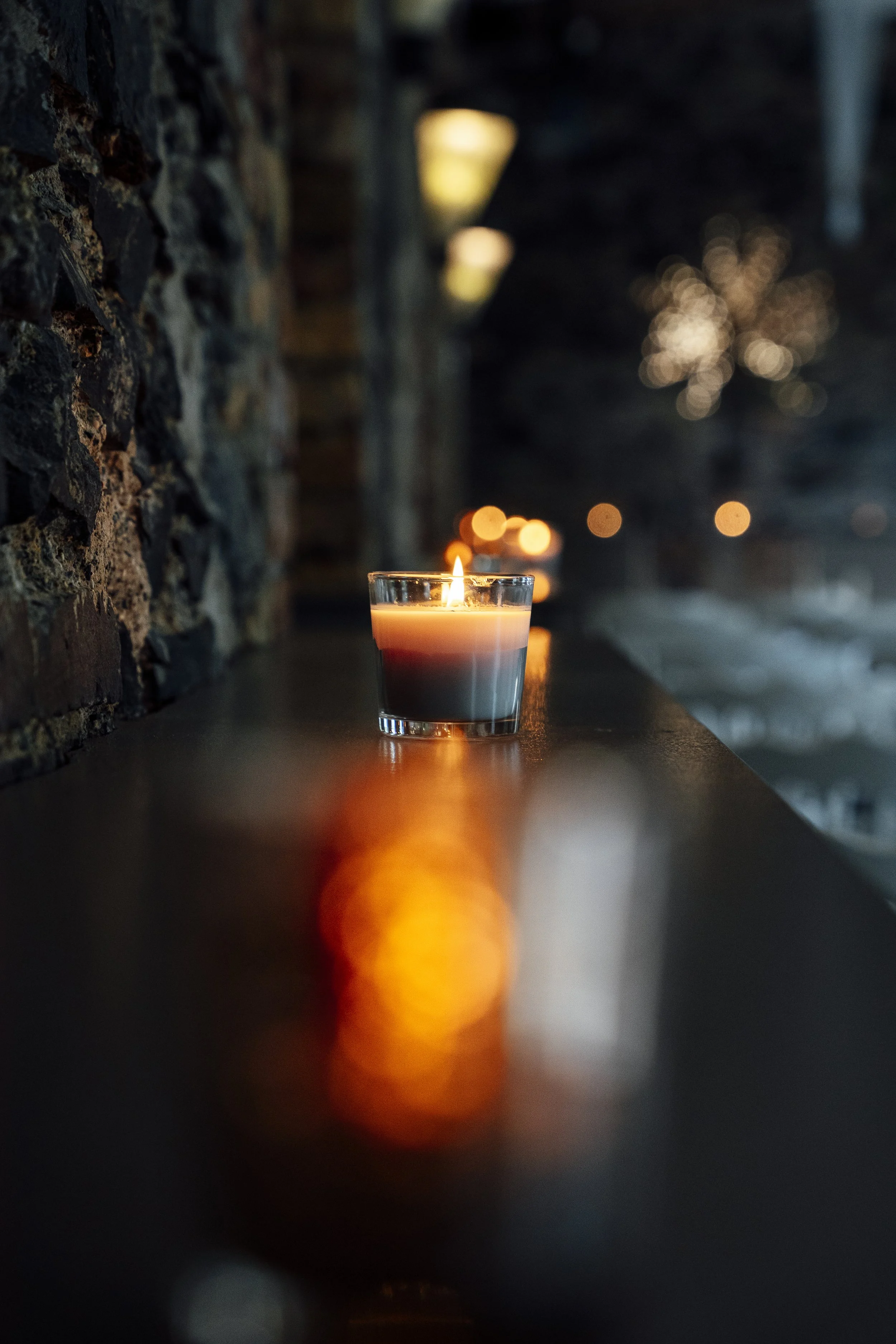 A lit candle in a glass holder on a dark surface, with a blurred background of lights and a stone wall. Yellow Brick Road Studio Wedding Photography