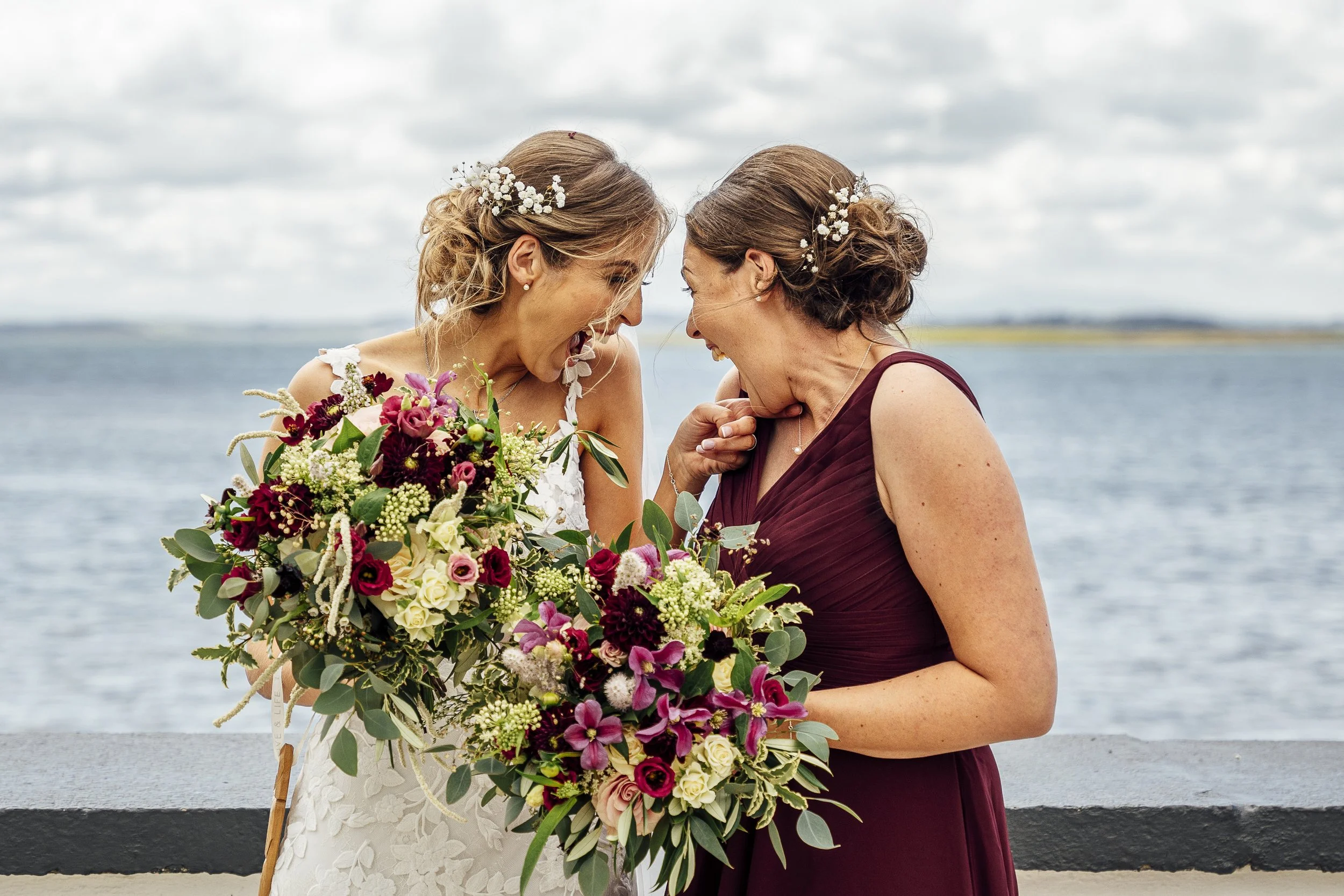 Two women celebrating a wedding, smiling and holding bouquets by the water. Yellow Brick Road Studio Wedding Photography