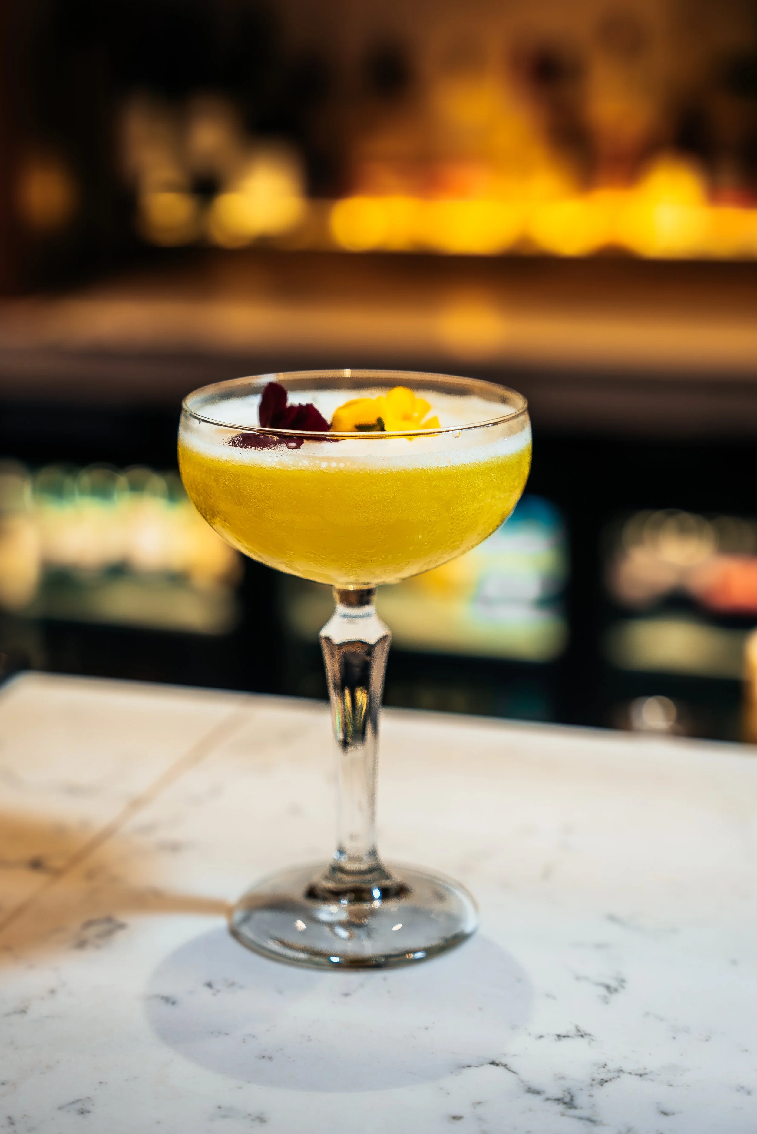 A yellow cocktail garnished with edible flowers on a white marble surface.