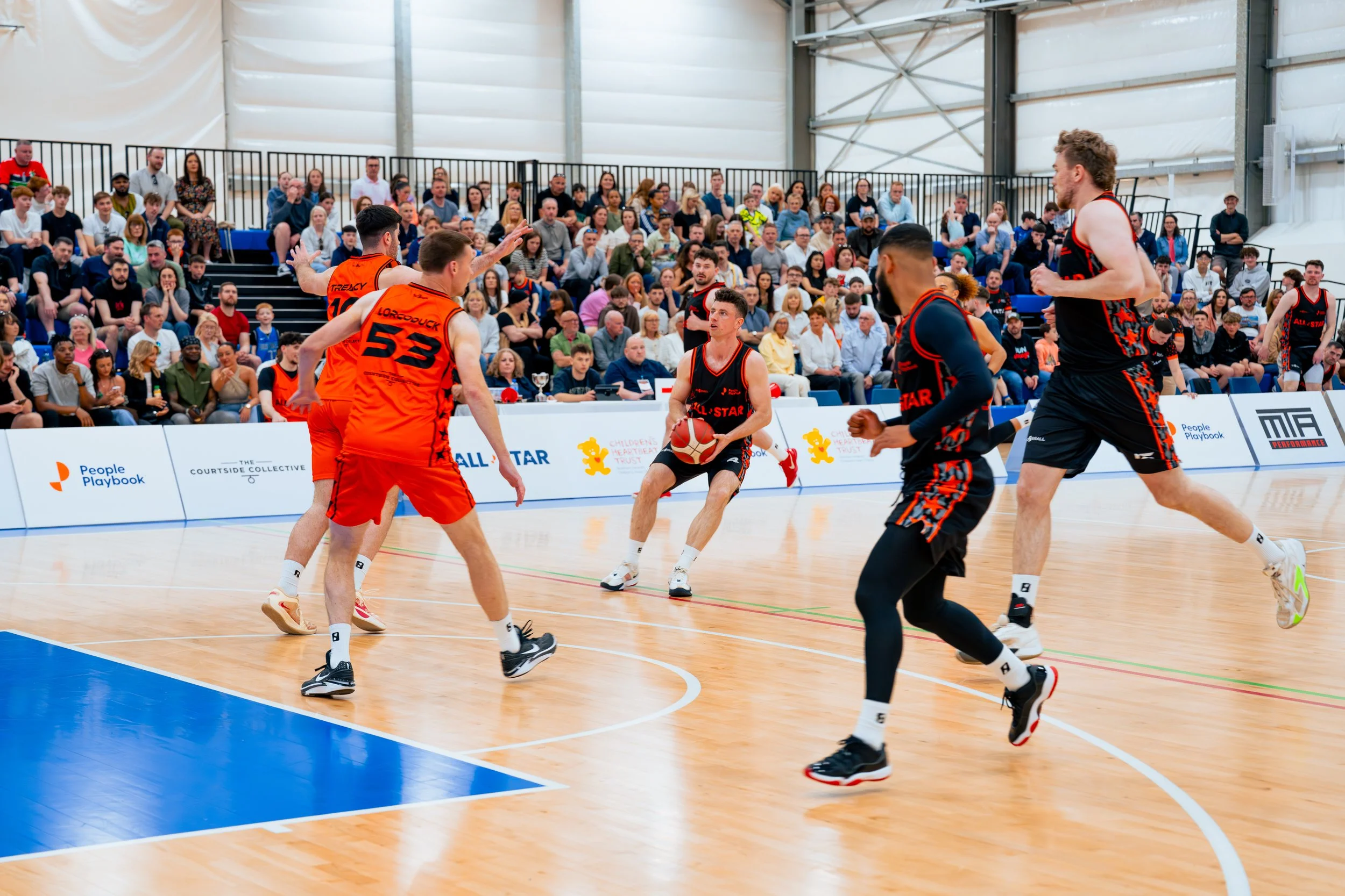 Basketball game with players in black and orange uniforms on an indoor court, and a crowd of spectators in the background. Courtside Collective Charity Basketball Game 