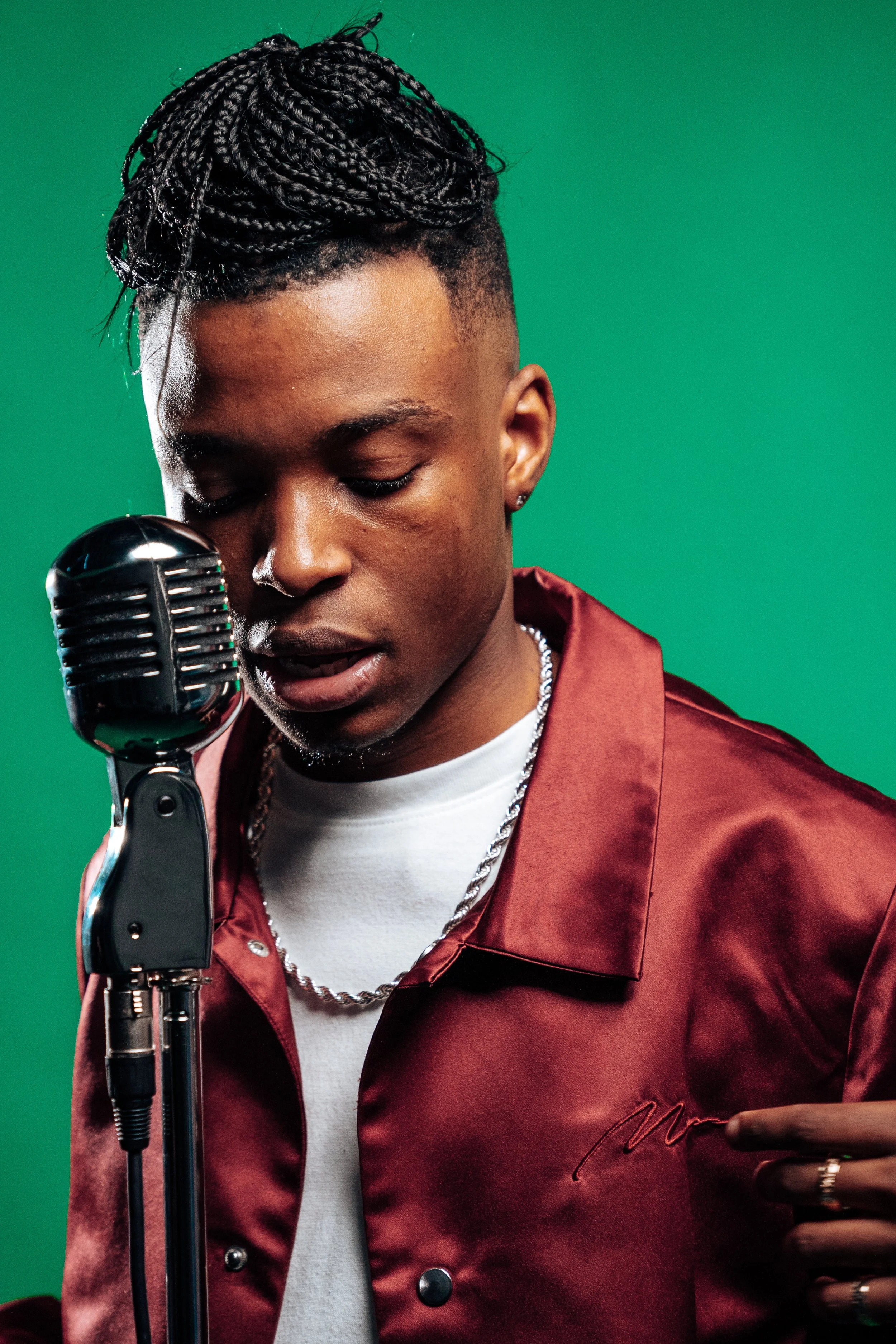 Young man with braided hair, wearing a satin jacket, silver chain, and white t-shirt, standing in front of a green screen, speaking or singing into a vintage microphone. Tanaka Belfast