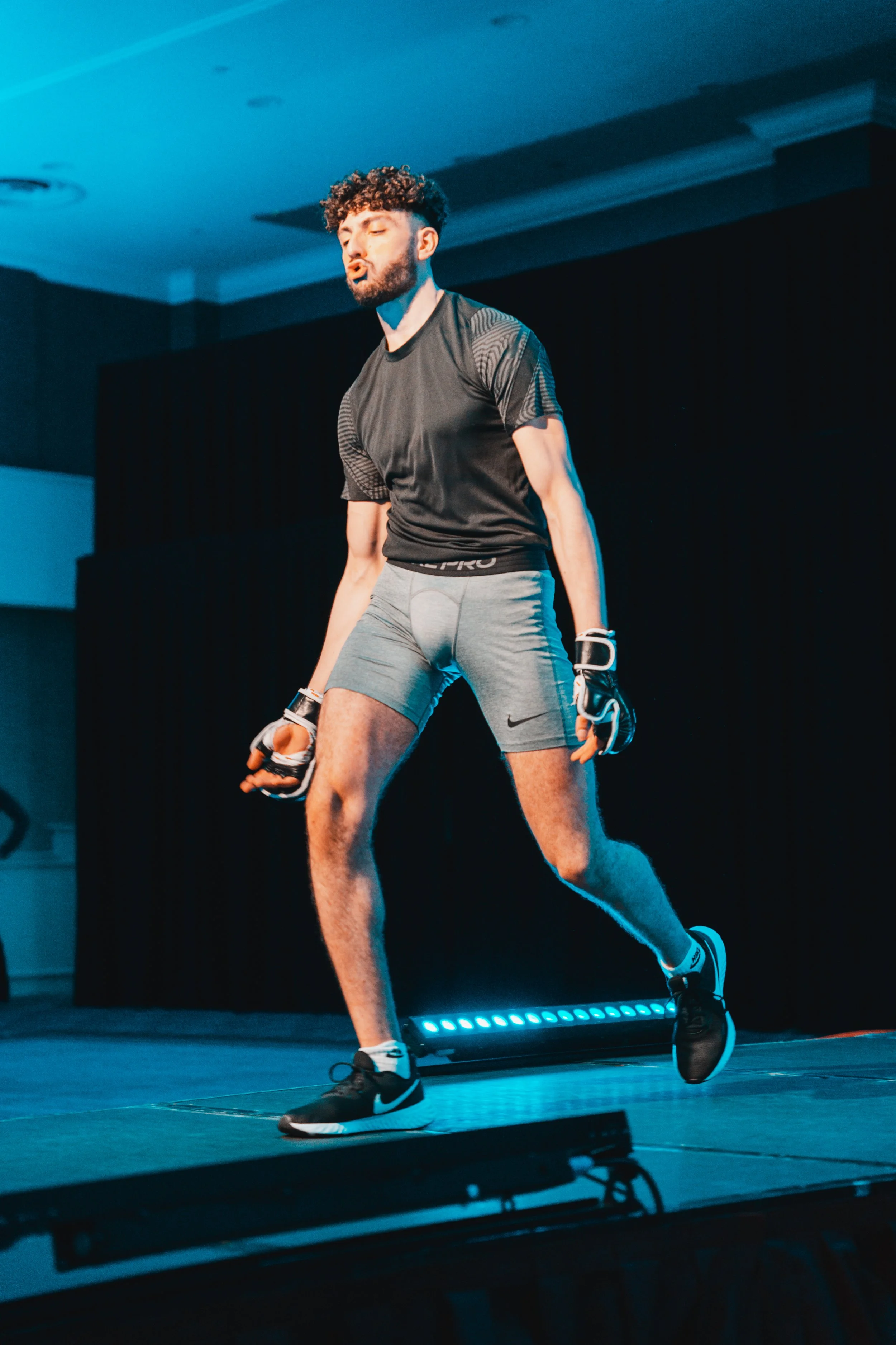 A male performer in athletic attire, including gloves, shorts, and sneakers, walks on a stage with a dark background and blue lighting.
