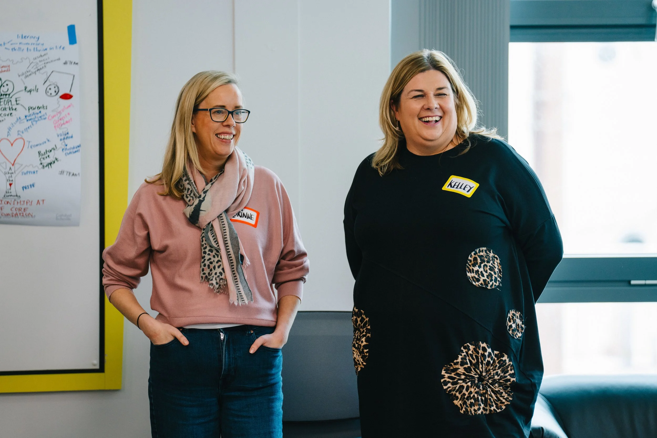 Two women smiling and standing indoors near a window. One woman wears glasses, a pink sweater, a printed scarf, and jeans, with a name tag that says 'BRINNE.' The other woman wears a black dress with leopard print circles and has a name tag that says