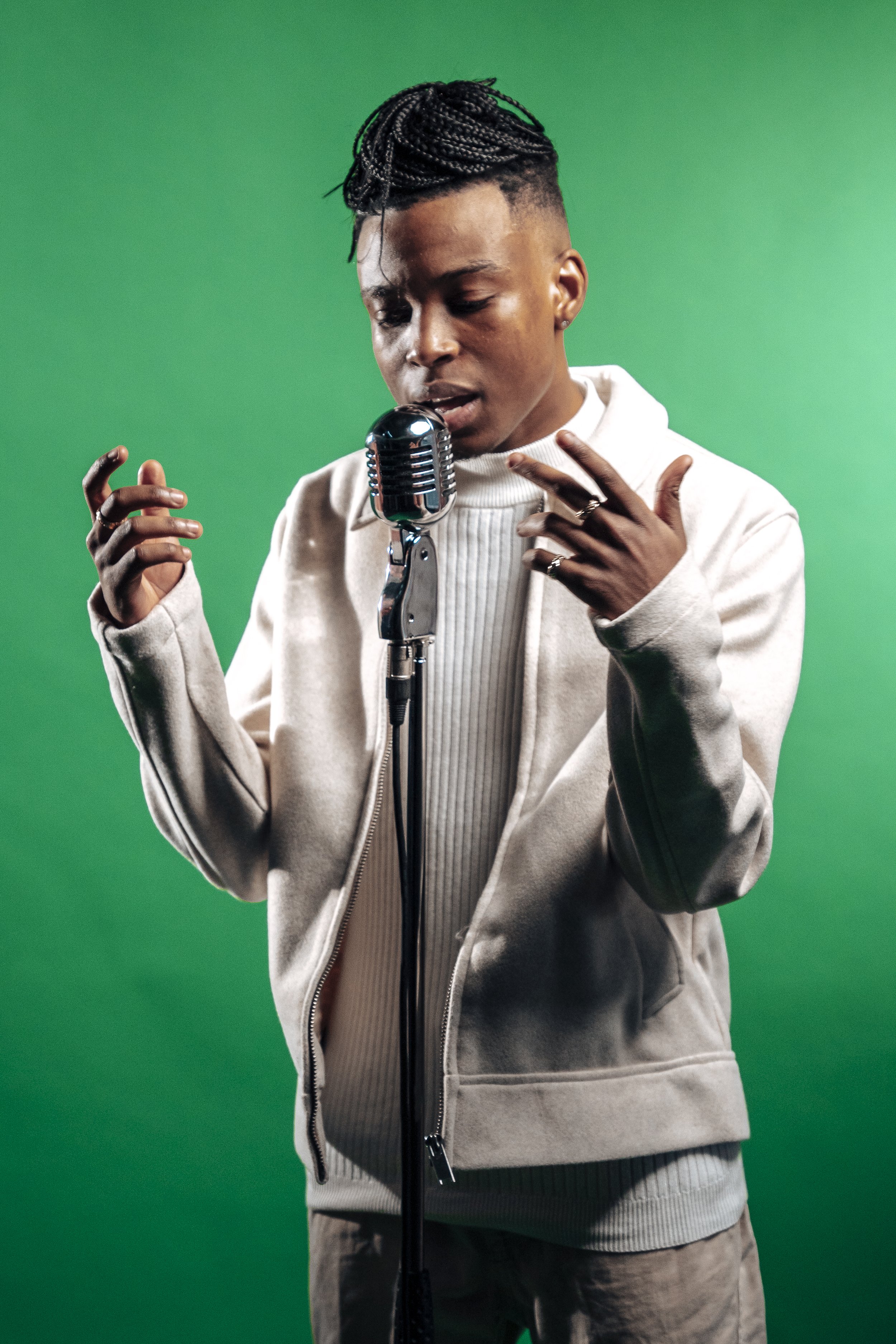 A person with braided hair singing into a vintage microphone on a green screen background. Tanaka Belfast