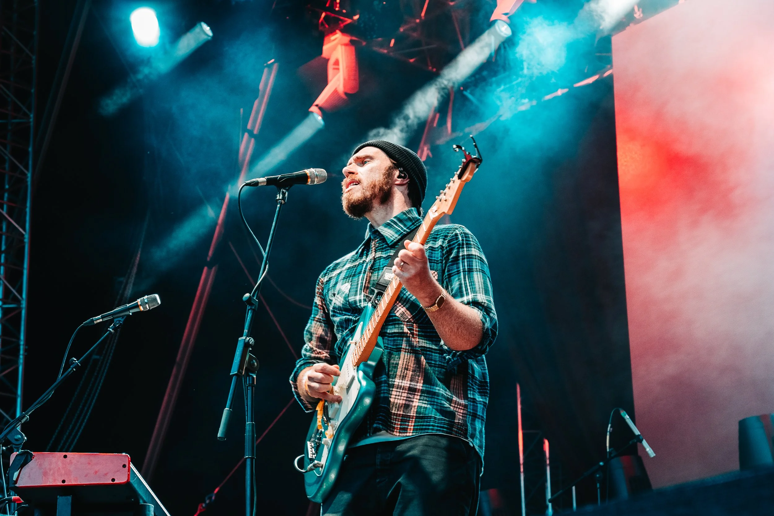 A male musician with a beard, wearing a black beanie and a blue checkered shirt, playing an electric guitar and singing into a microphone on stage with colorful stage lights and fog effects.